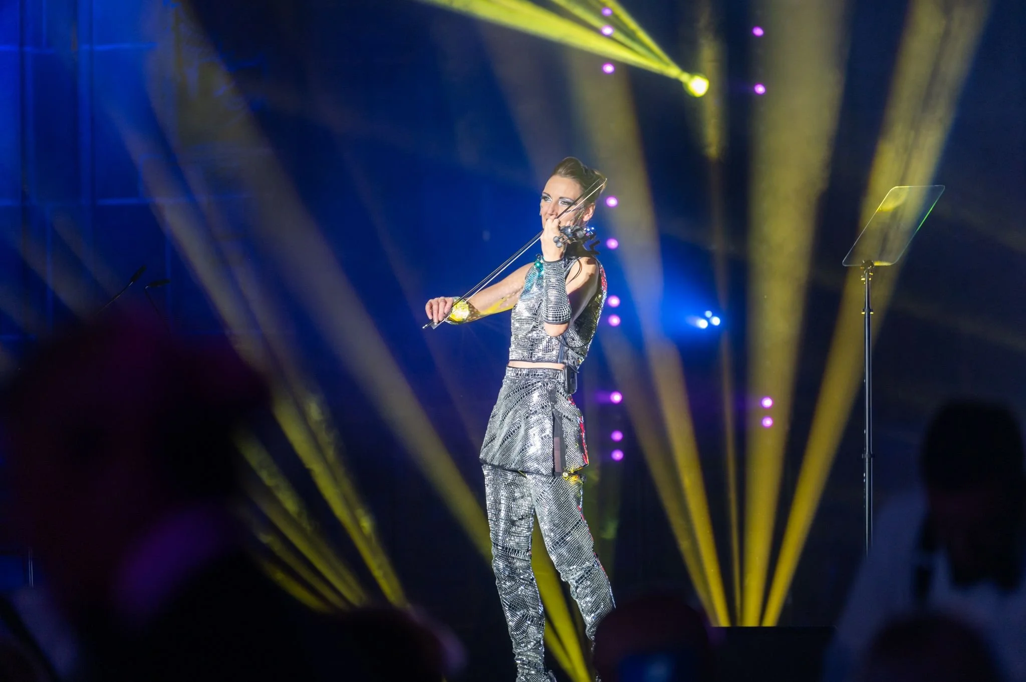 A woman dressed in a shiny silver outfit playing the violin on stage under yellow and purple stage lights.