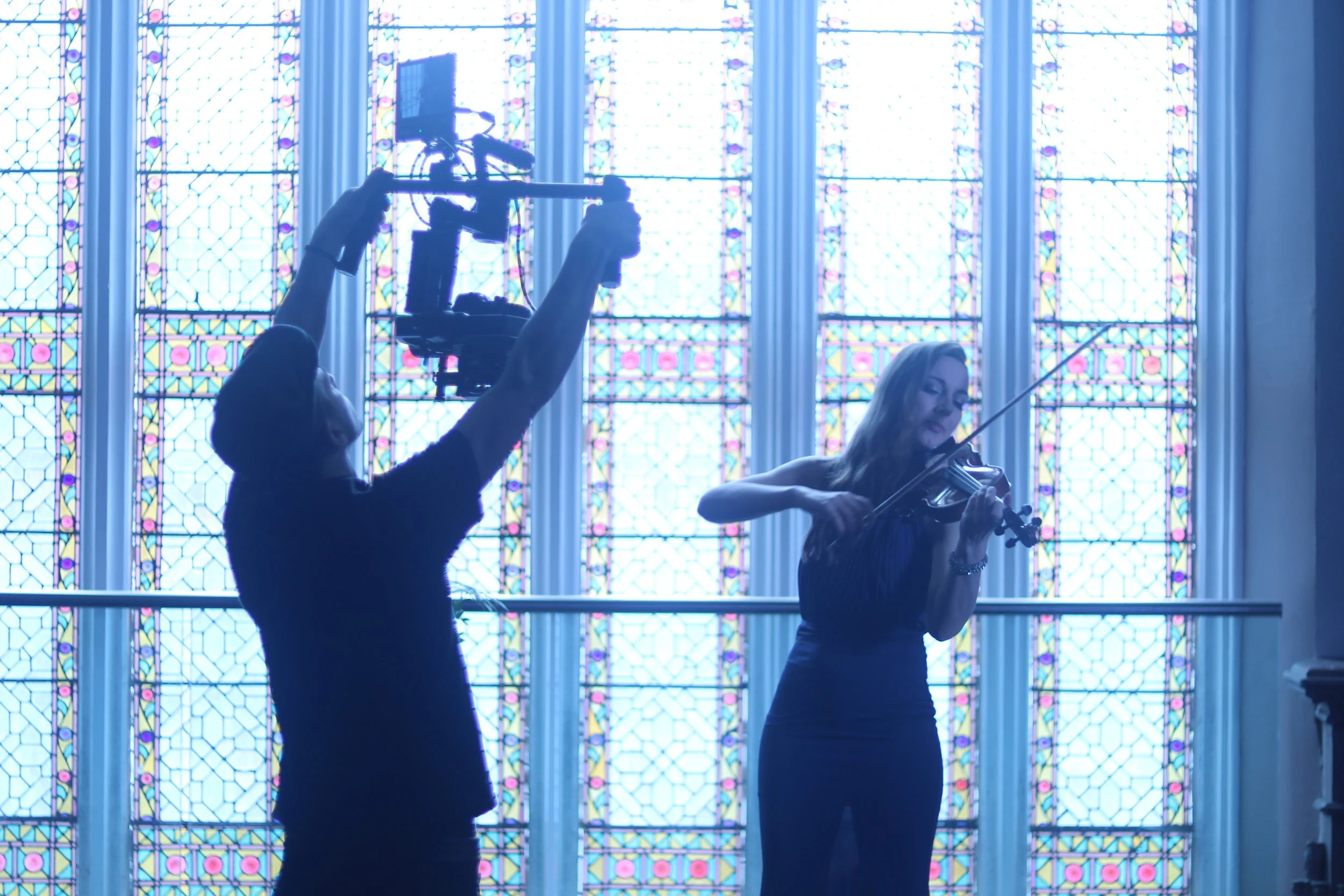 Two women playing violins in front of stained glass windows, with one woman holding a camera mounted on a stabilizer.