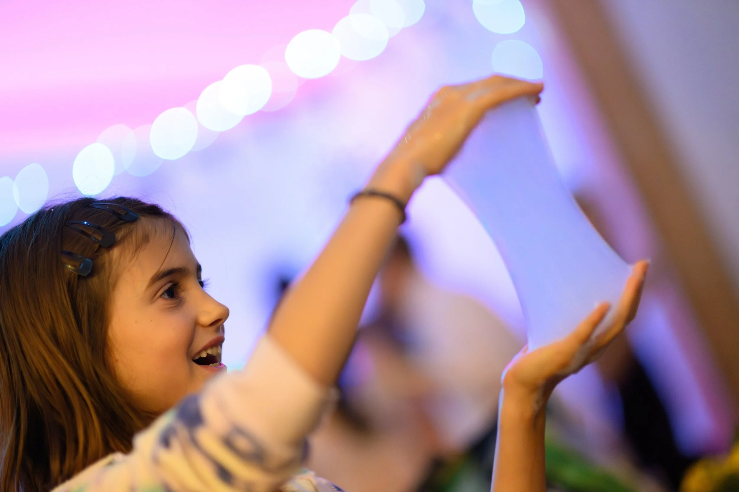 A young girl with long hair and hair clips is smiling and reaching up, likely in a playful or joyful moment, against a colorful background with pink and purple bokeh lights.