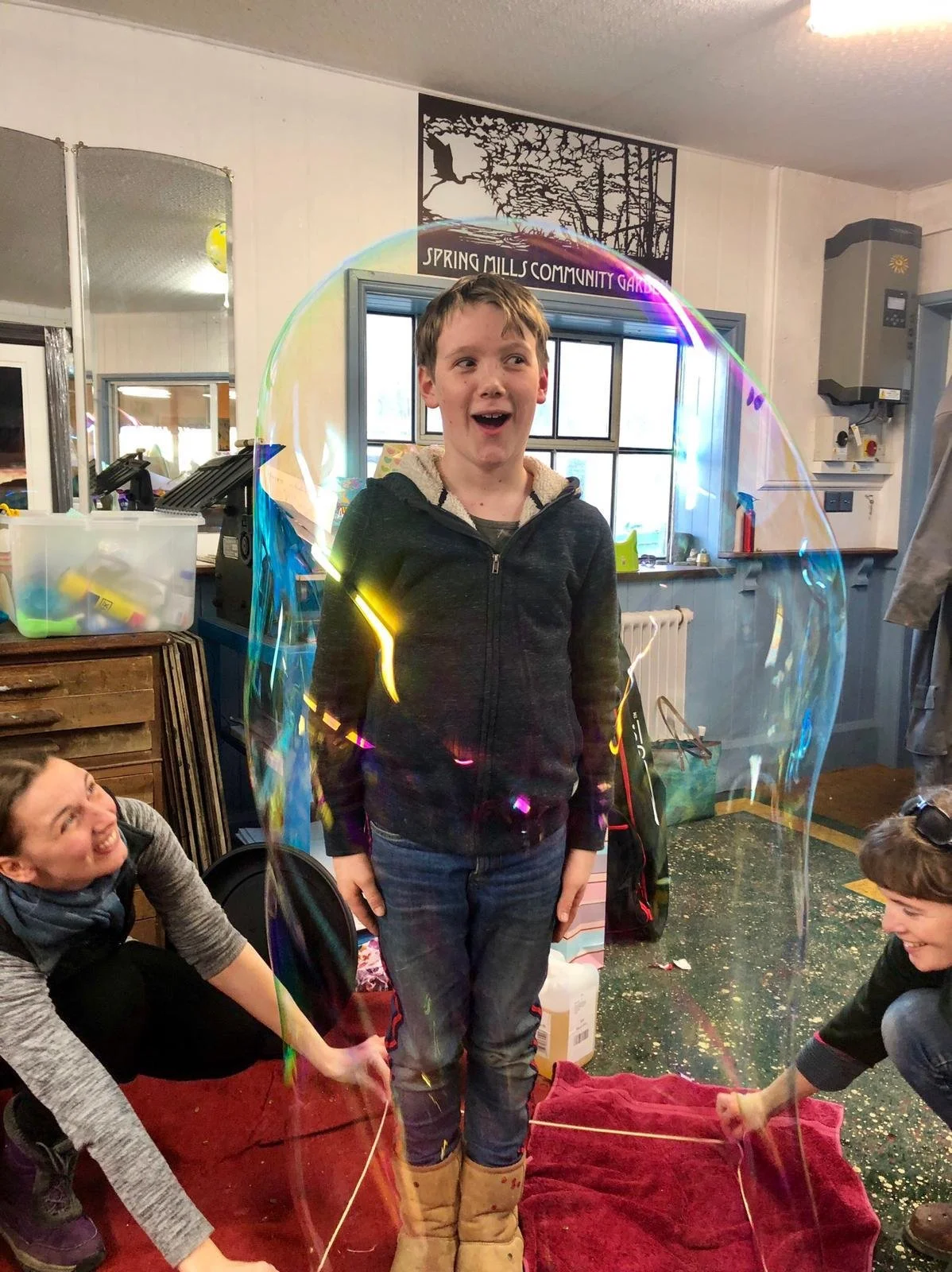 A boy stands inside a large soap bubble with three children on the floor around him, one girl smiling and holding a string attached to the bubble, in a room with a sign reading 'Spring Mills Community' on the wall.