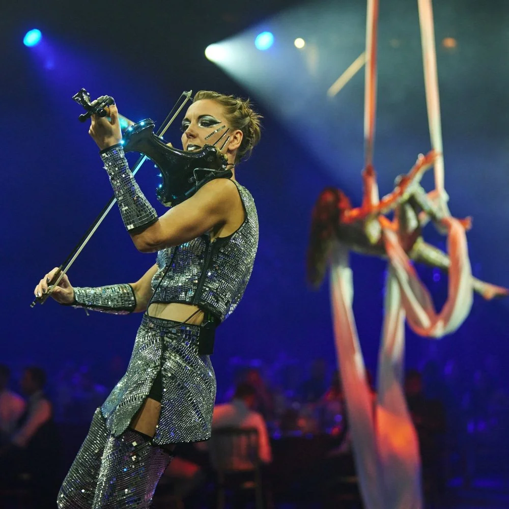 Performer in a silver, sequined outfit playing a black electric violin with dramatic makeup at a concert, with colorful stage lights in the background.