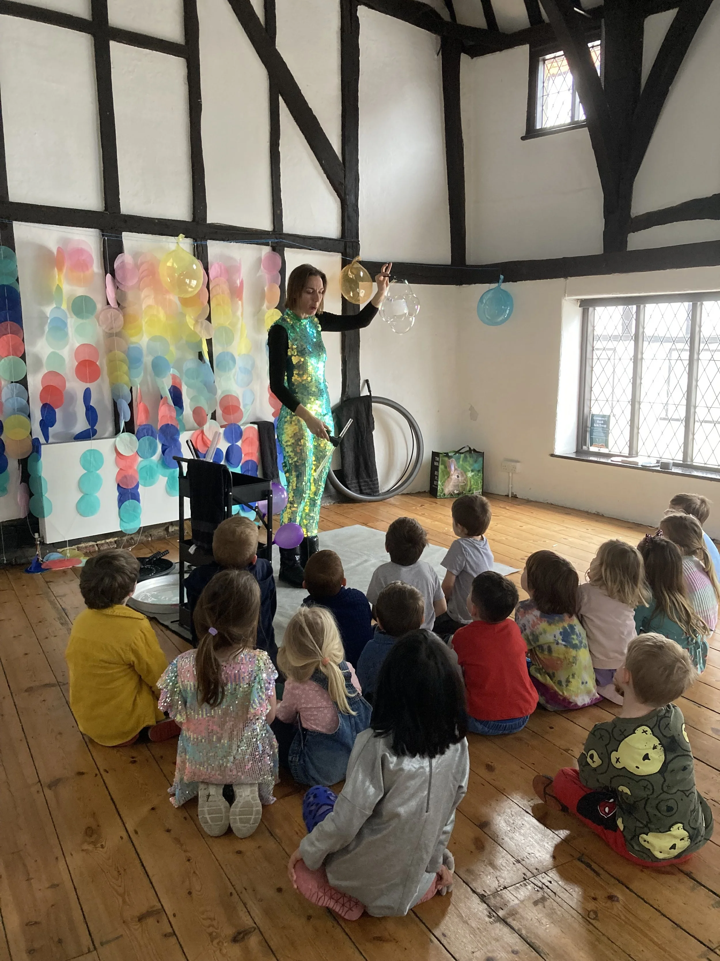 A woman dressed in a shiny, colorful costume performing magic or entertaining in front of a group of children sitting on a wooden floor. The background includes balloons and a decorative curtain. The scene is inside a room with a high ceiling and woo