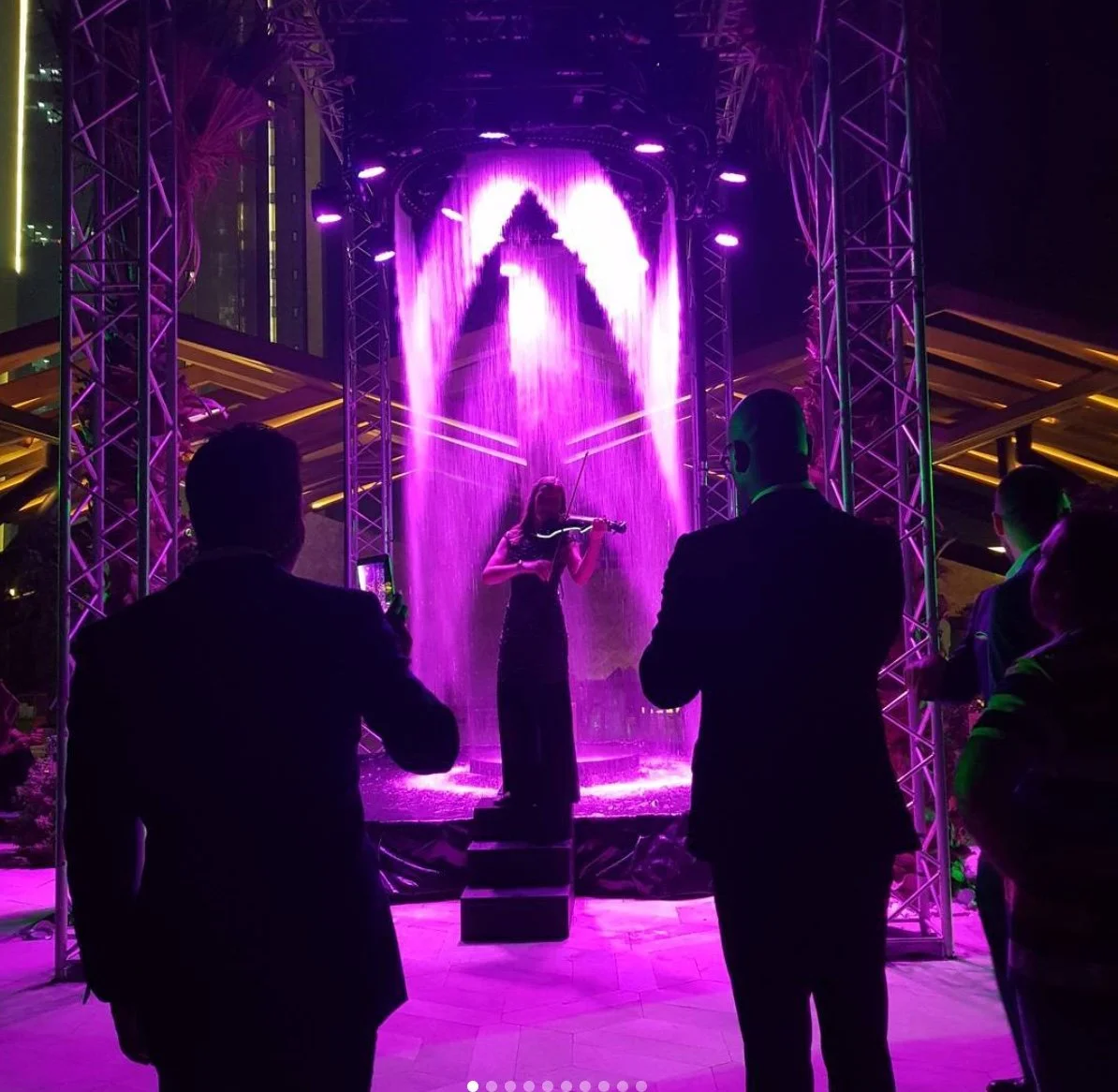 A woman playing the violin on a stage with purple water fountain lights, surrounded by silhouetted audience members in formal attire at night.