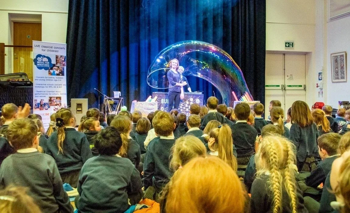Children seated on the floor watching a performer creating a large soap bubble on a stage in a school auditorium.