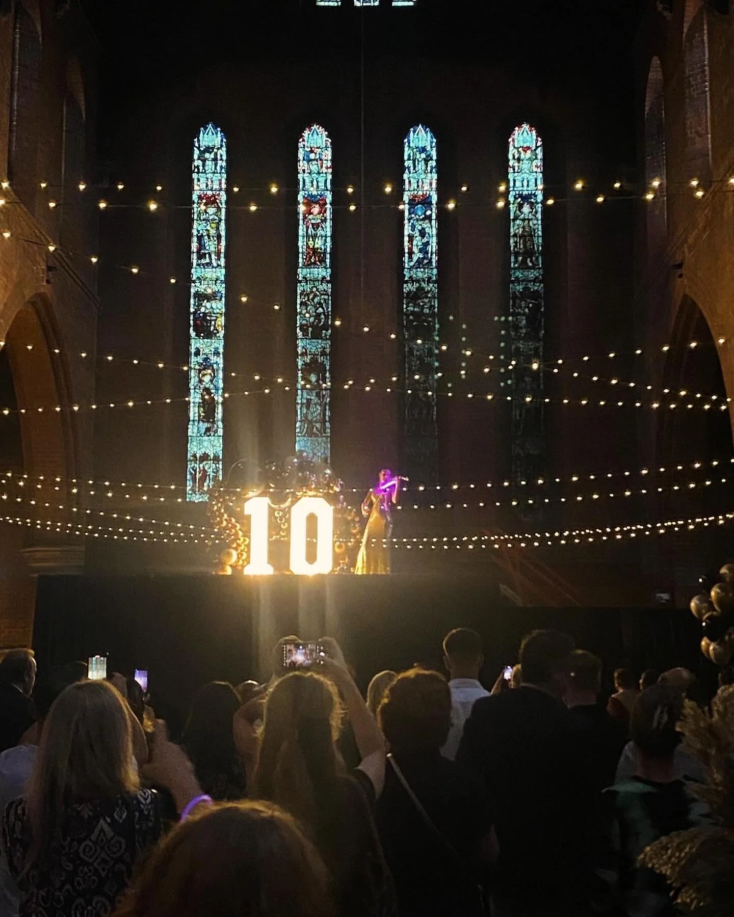 A large crowd gathered in a church, celebrating a 10th anniversary. The church has tall stained glass windows and string lights hanging from the ceiling. There is a stage with a large illuminated number 10, balloons, and a performer singing or speaki