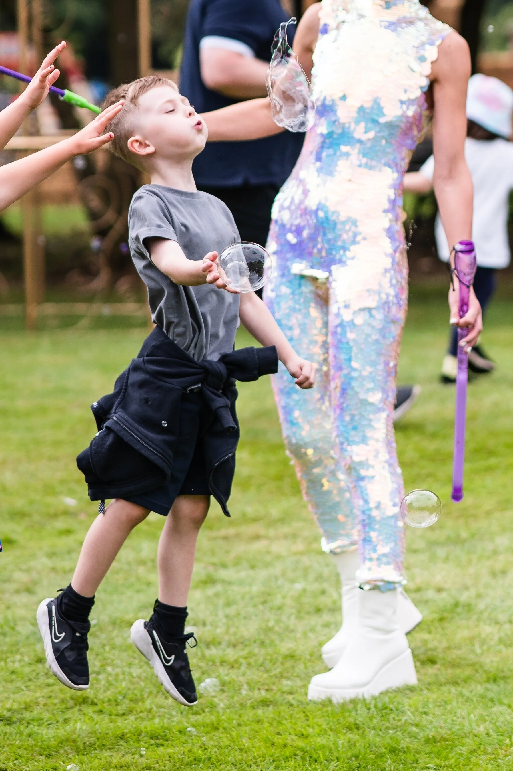 A young boy with blond hair blowing bubbles at an outdoor event, wearing a grey t-shirt, black shorts, and black Nike sneakers, with a jacket tied around his waist. A woman in a colorful, sequined dress stands nearby, possibly helping with the bubble