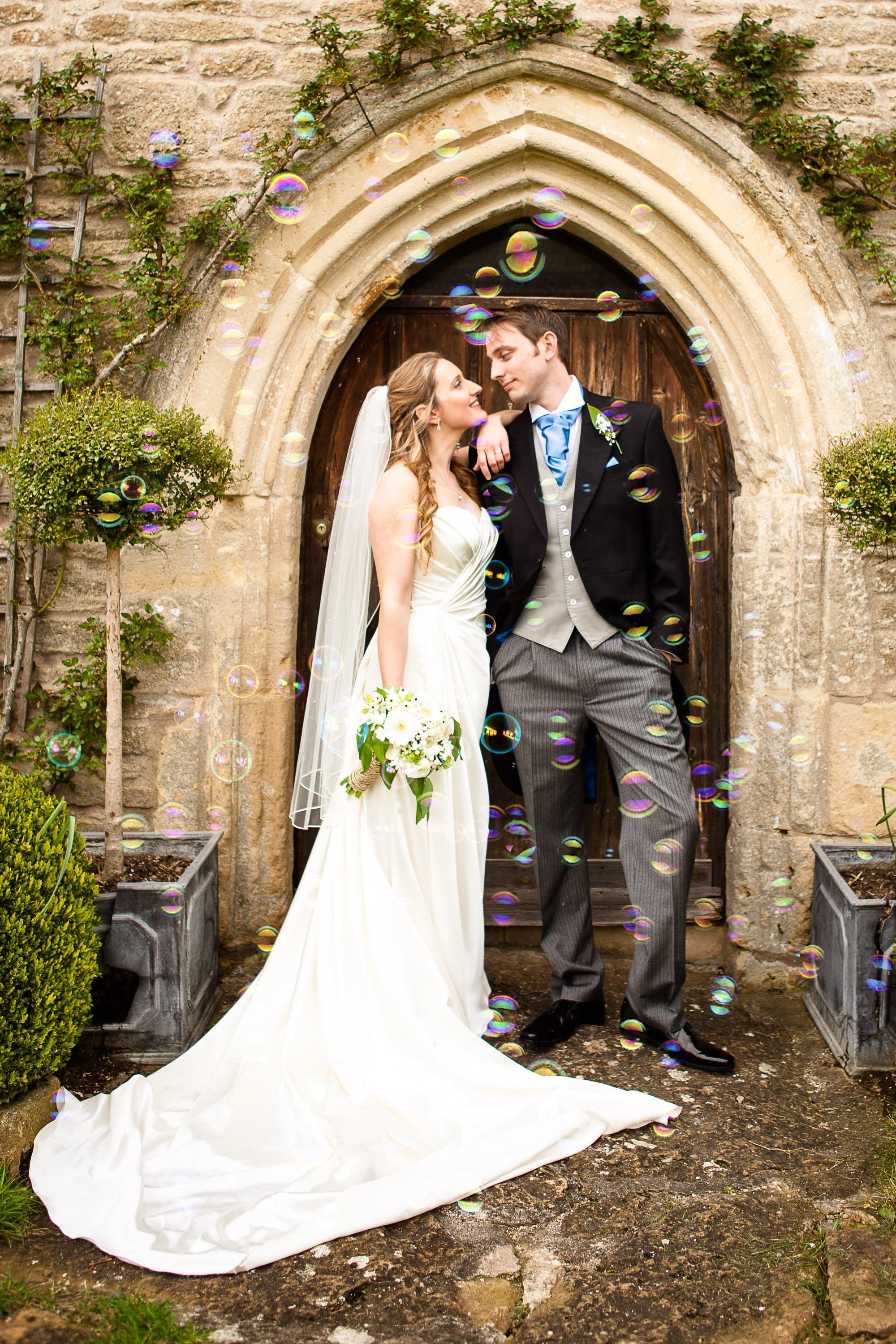Bride and groom standing together outside a stone archway, surrounded by greenery, with soap bubbles floating around them during a wedding celebration.
