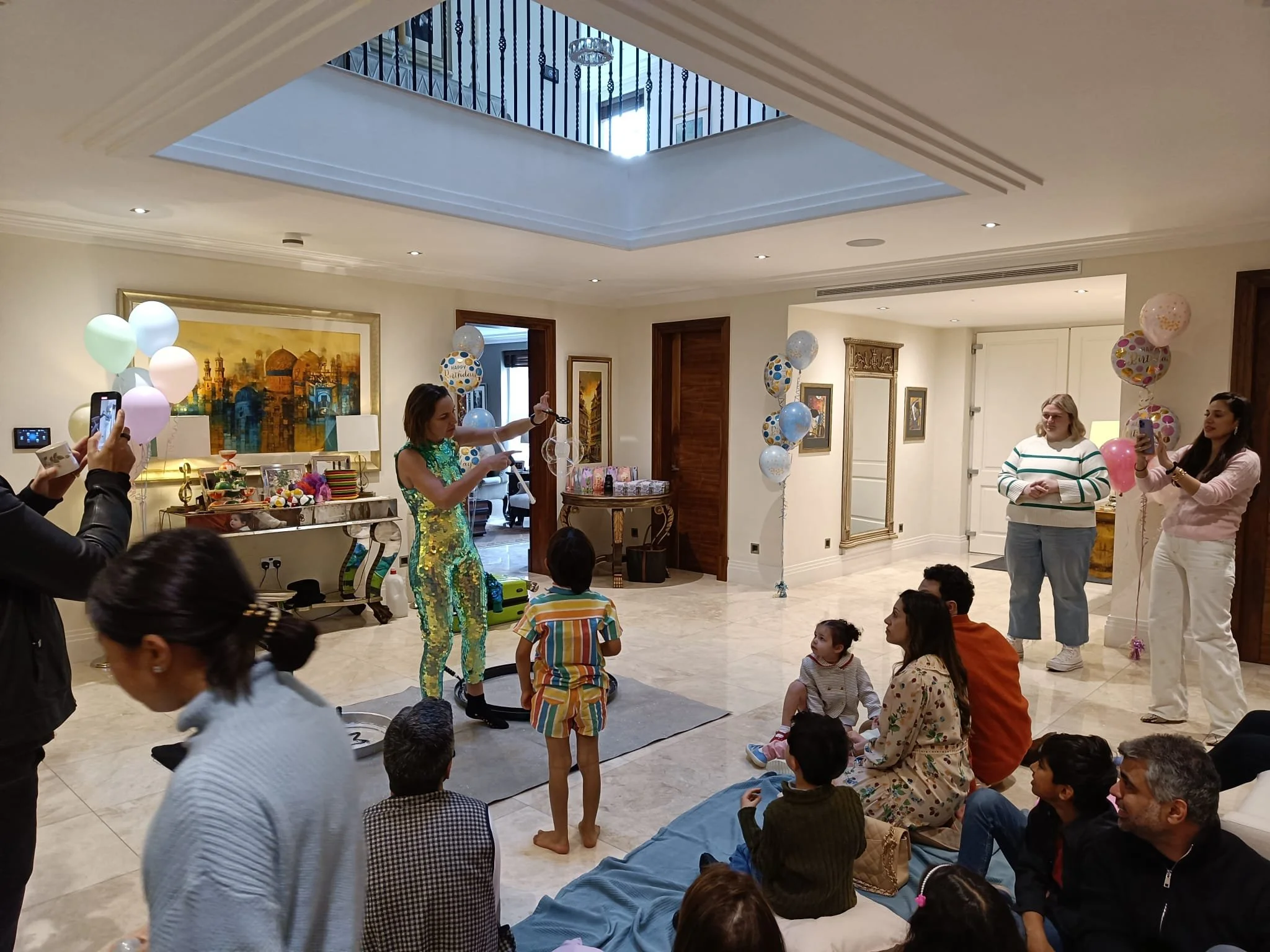 A birthday celebration in a spacious room with a crowd of children and adults. There are balloons, a performer or entertainer in a shiny outfit, and some adults taking pictures or videos. The room has artwork on the walls and a skylight ceiling.