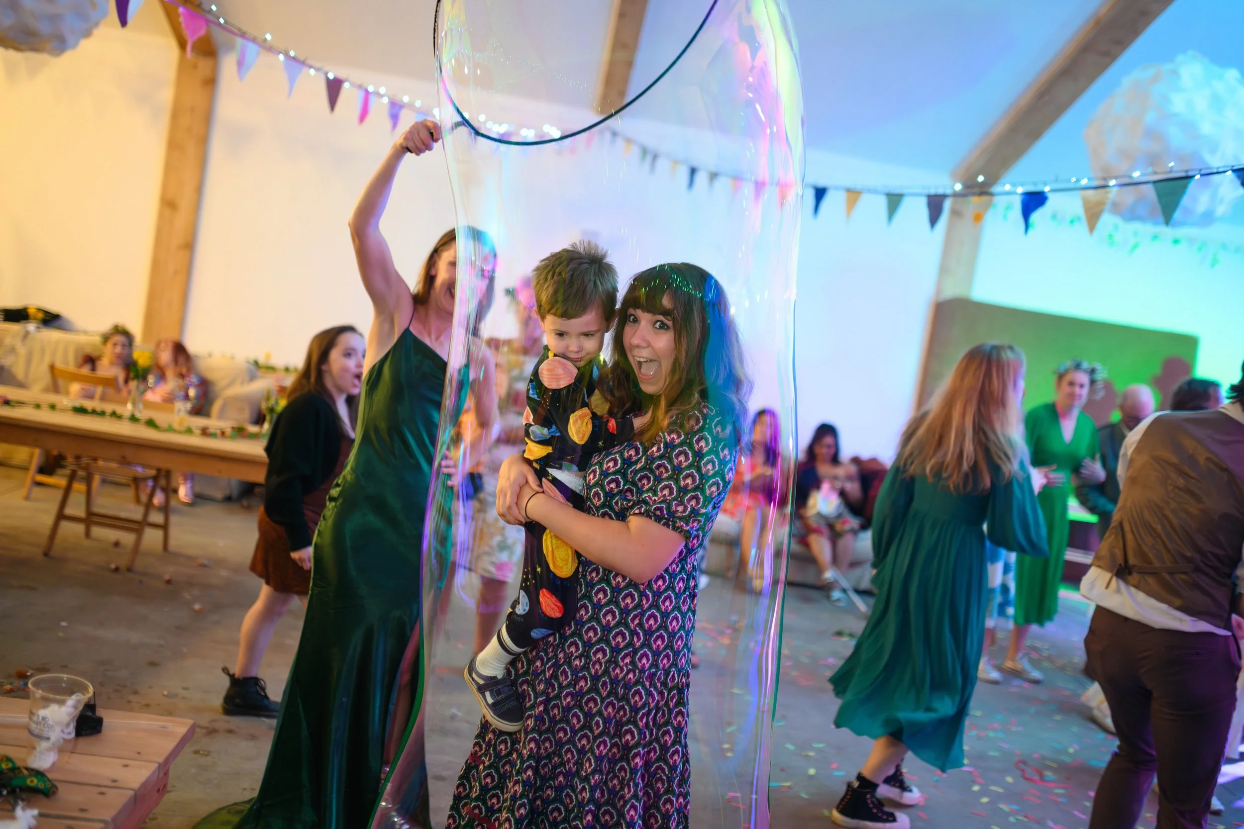 People at a party dance and play with a large bubble inside a decorated hall with string lights and colorful banners.