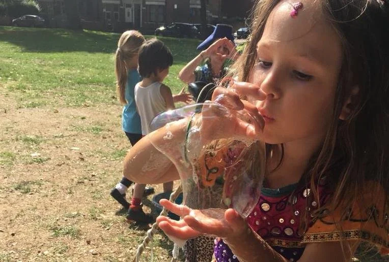 A young girl is blowing bubbles outdoors in a park, with other children in the background enjoying the day.