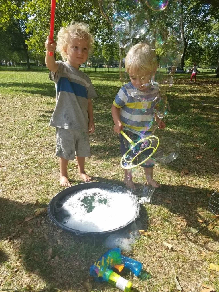 Two young boys playing with soap bubbles outdoors in a park with green trees in the background. One boy holds a bubble wand, and the other is using a bubble ring. A tray of soap solution and bubble-blowing toys are on the ground.
