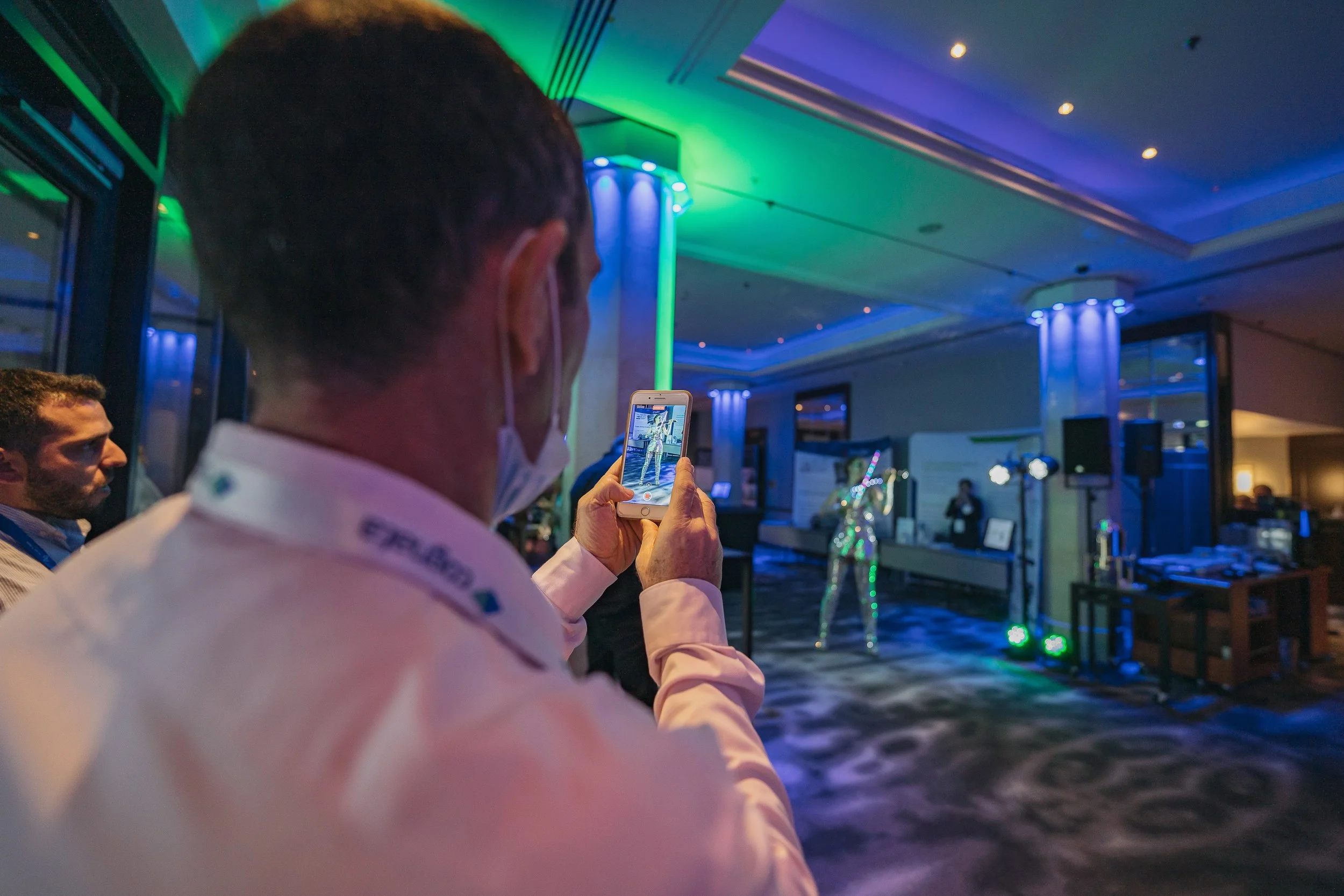 A person wearing a pink shirt and face mask is taking a photo or recording on a smartphone, at an event with colorful lighting, in a large indoor space with people and decorations.