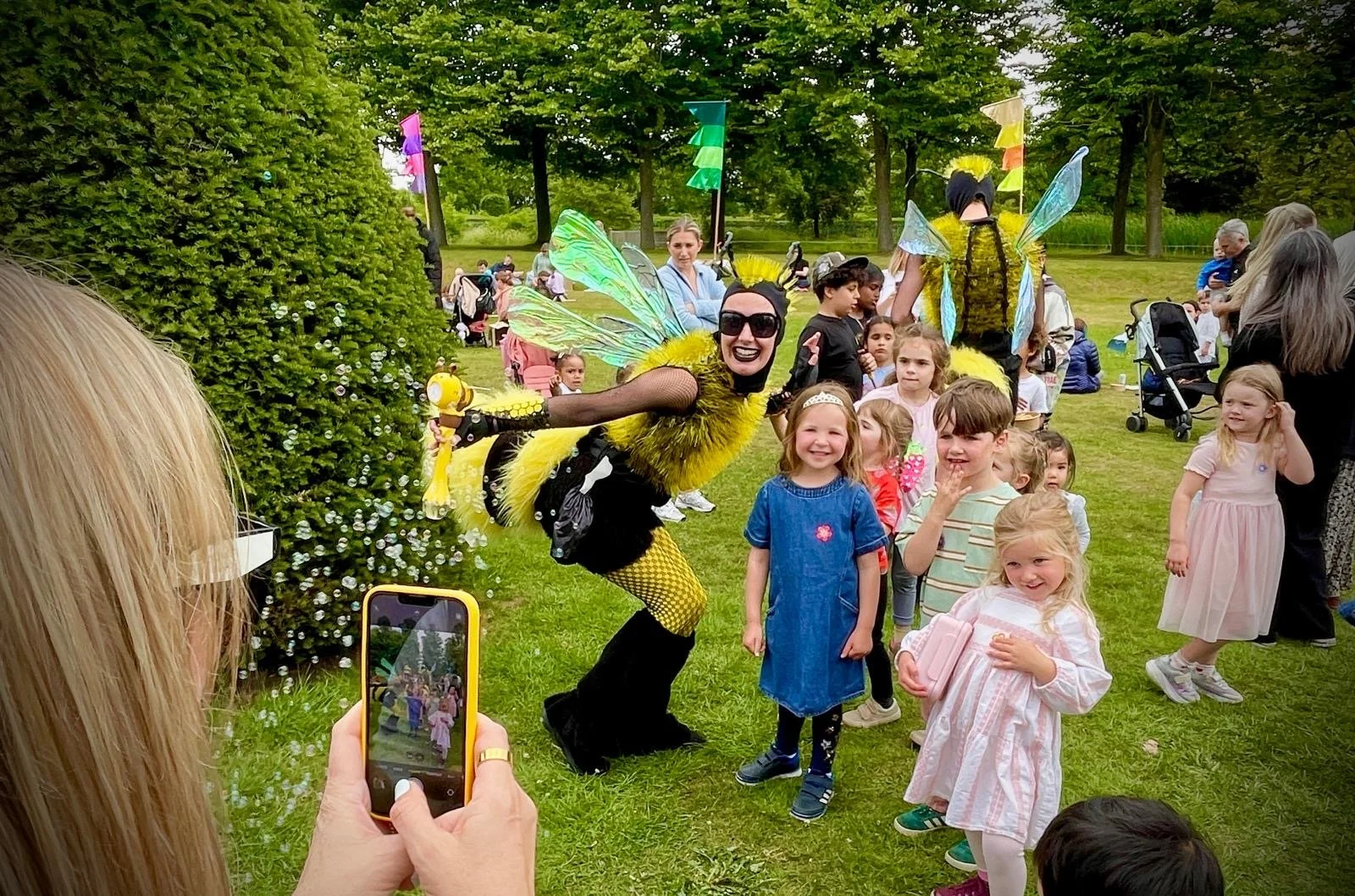 A group of children and adults gathered outdoors in a park, taking photos and watching as two performers dressed as colorful bees entertain the crowd. The performers wear costumes with large yellow and black bodies, iridescent wings, and yellow pom-pom accents, while the children look on smiling. Lush green trees and grass surround the scene.