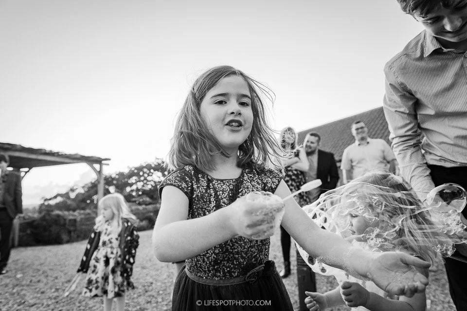 A young girl making bubbles outdoors with other children and adults in the background.