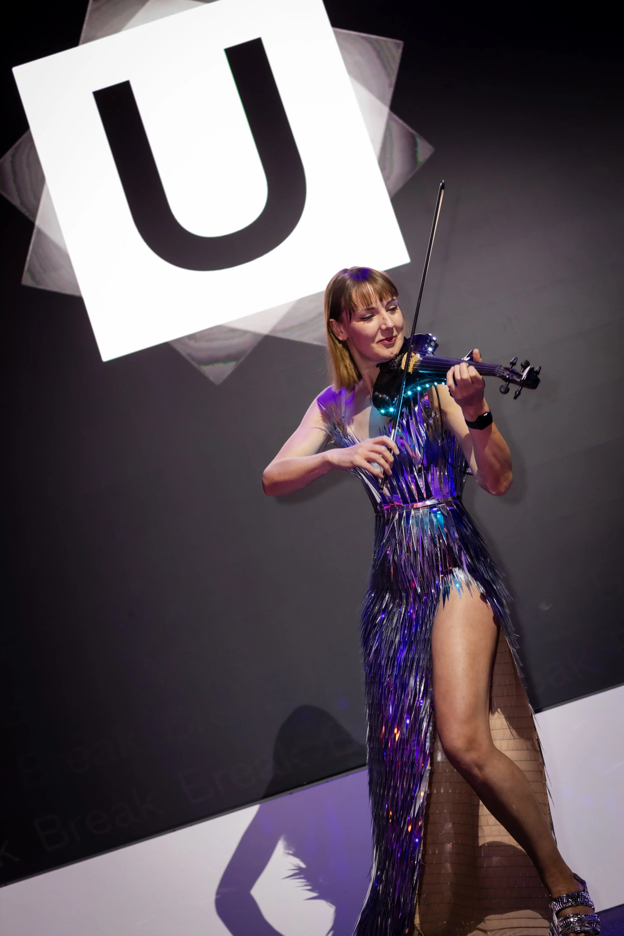 A woman playing a violin in a shimmering, silver dress with a slit, standing in front of a large illuminated sign with a bold black letter 'U' on a white background.