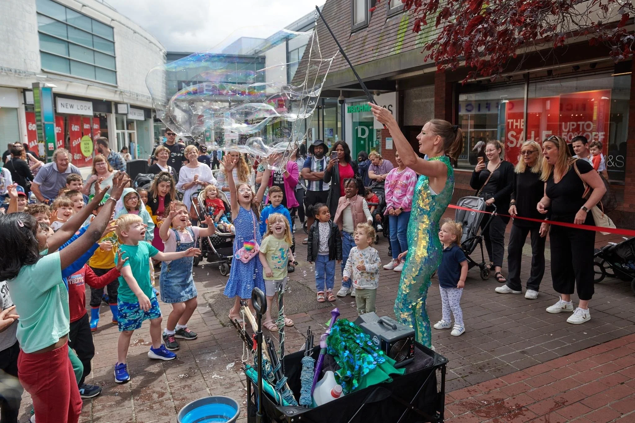 A woman performing a bubble show for children and adults on a busy city street, with spectators watching and taking photos.