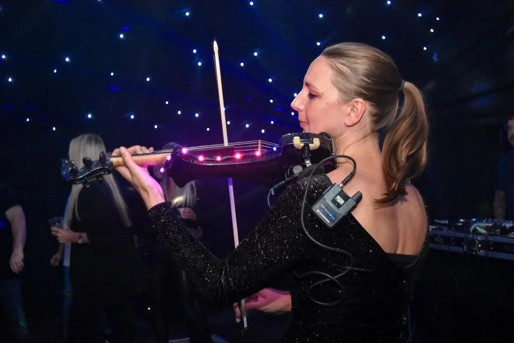 A woman playing a violin with LED lights on the neck, wearing a black sparkly dress at a party or concert, with star-like lights in the background.