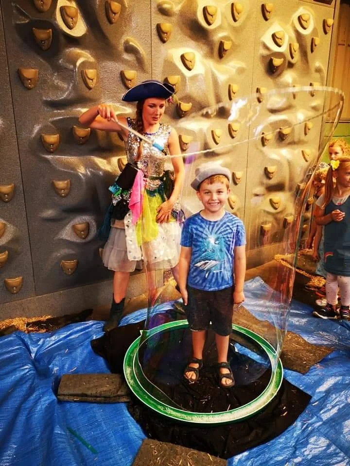 A young boy standing inside a large soap bubble with a smiling woman in a pirate costume nearby, next to a climbing wall at an indoor activity center.