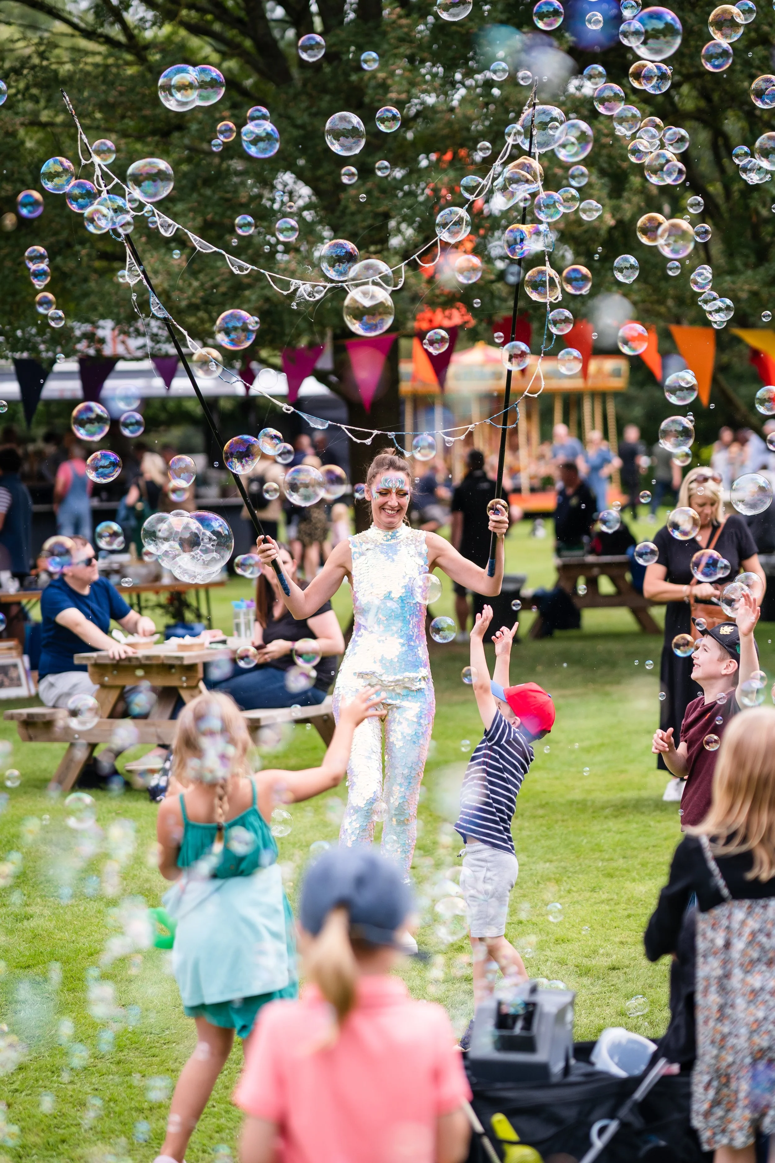 A woman with face paint and a shiny dress is entertaining children with bubbles at an outdoor party. People are seated at picnic tables, and colorful banners are hung in the background.