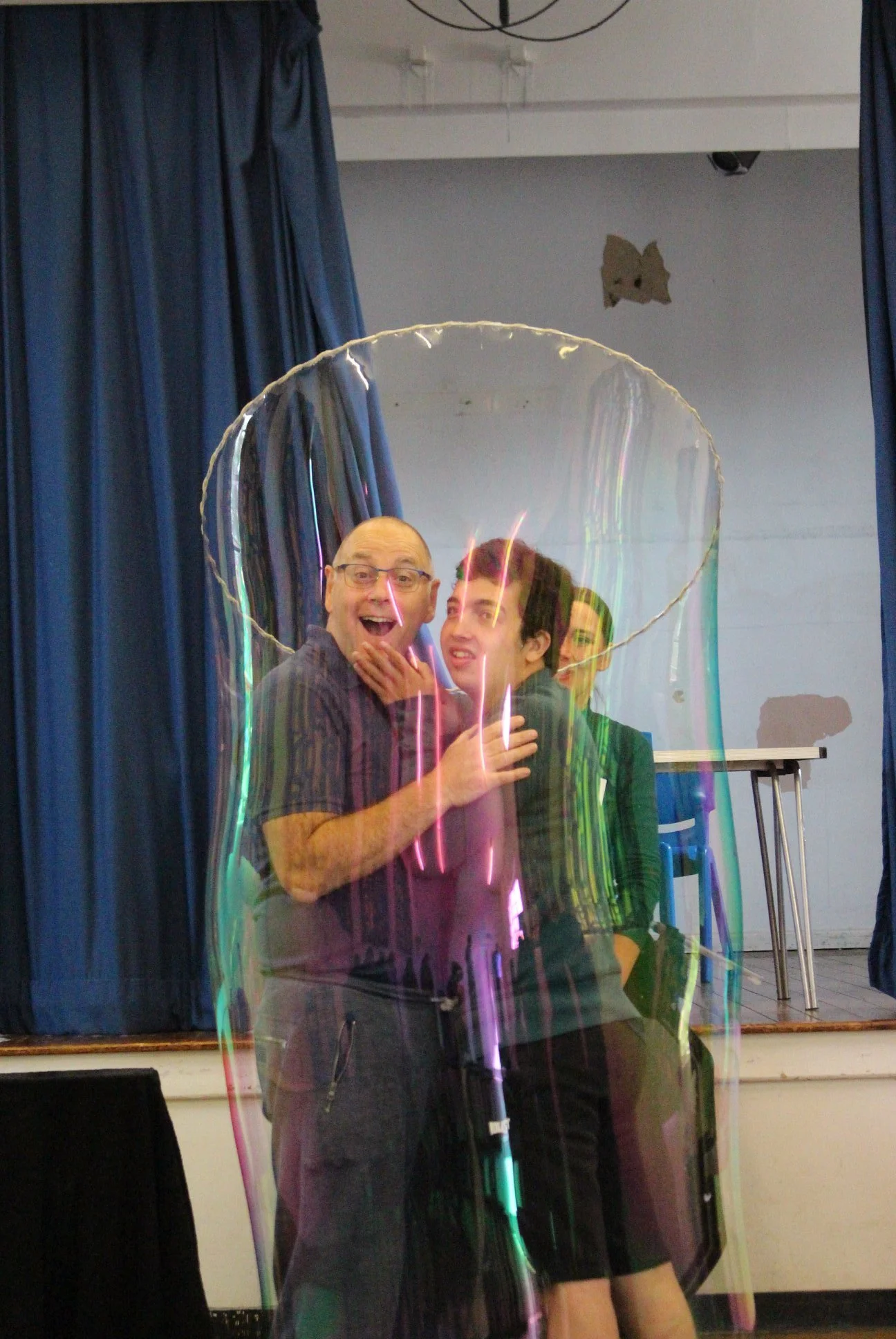 Two men and one woman are taking a selfie in front of a mirror, with colorful reflections. They are smiling and standing close together, with the man on the left holding the phone.