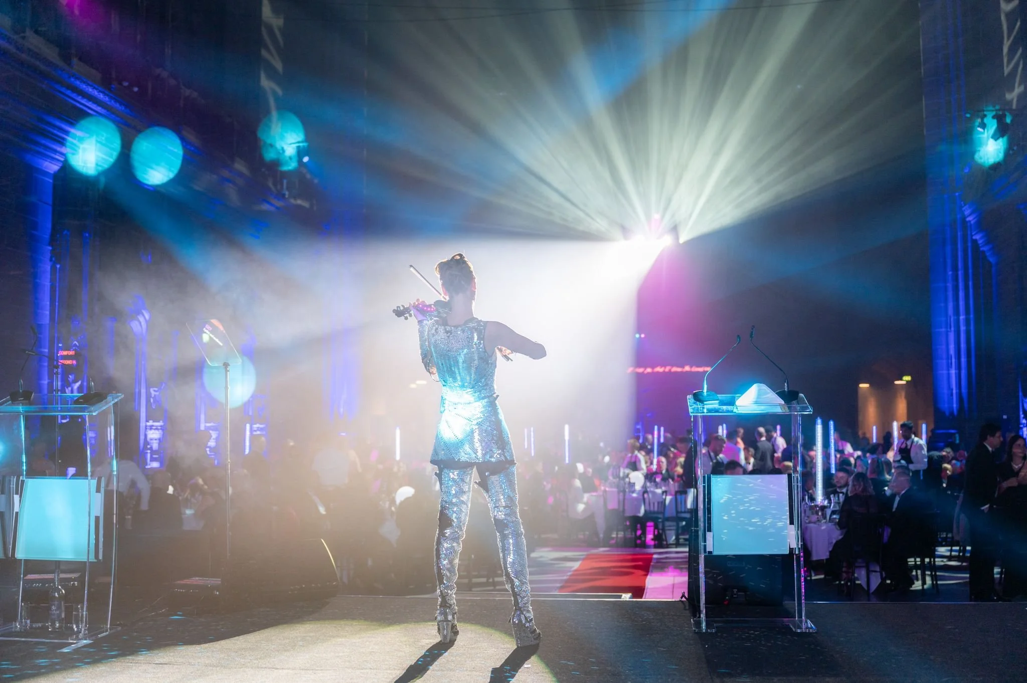 A woman dressed in sparkly silver attire performing with a violin on a stage at a lively event, with colorful lights and an audience in the background.