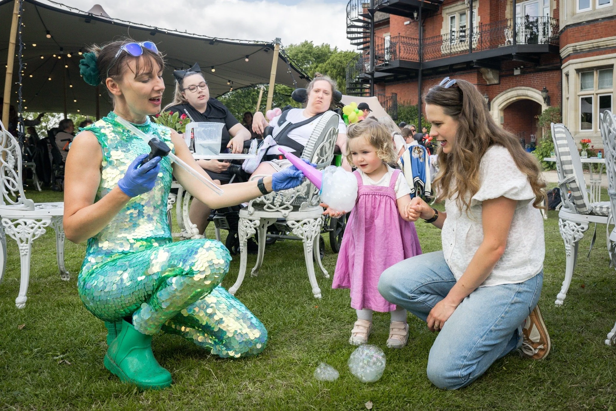 A woman dressed as a mermaid with a shiny, sea-green costume kneels on the grass and shows a young girl a cotton candy during an outdoor event with several people in the background.