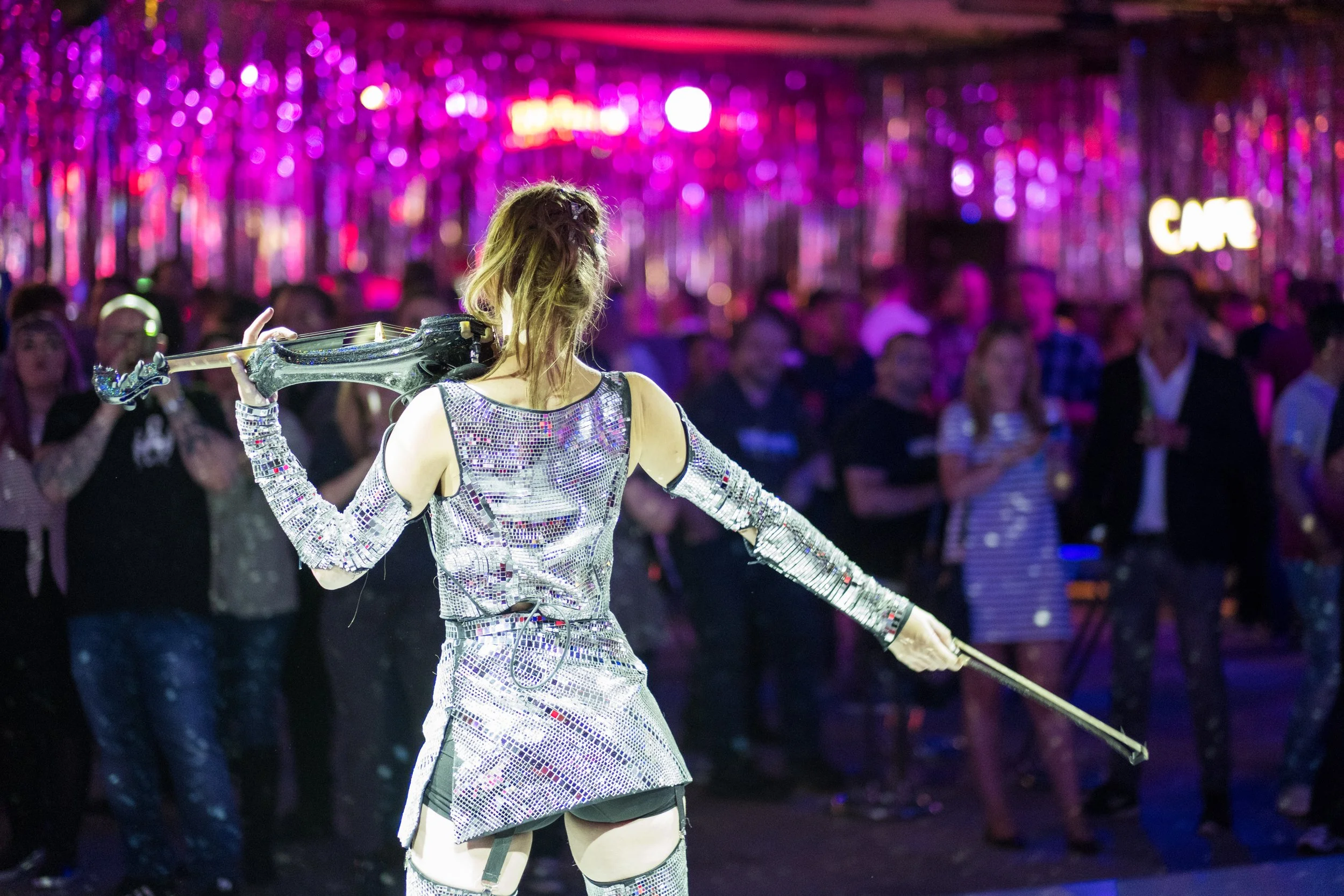 A performer in a glittery silver costume playing a violin in front of a colorful, blurry crowd at a nightclub or concert.