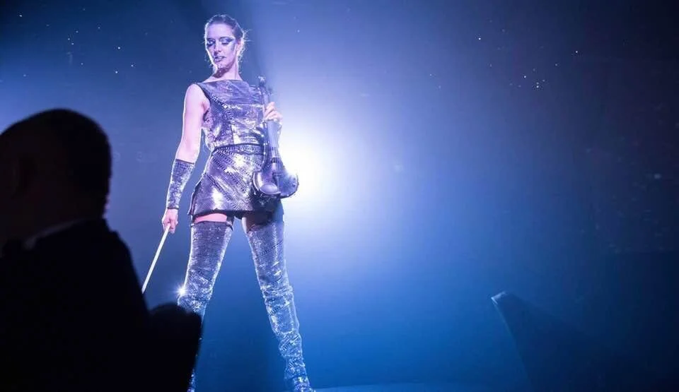 A woman in a shiny, metallic outfit with matching gloves and thigh-high boots, standing on a stage with a spotlight behind her.
