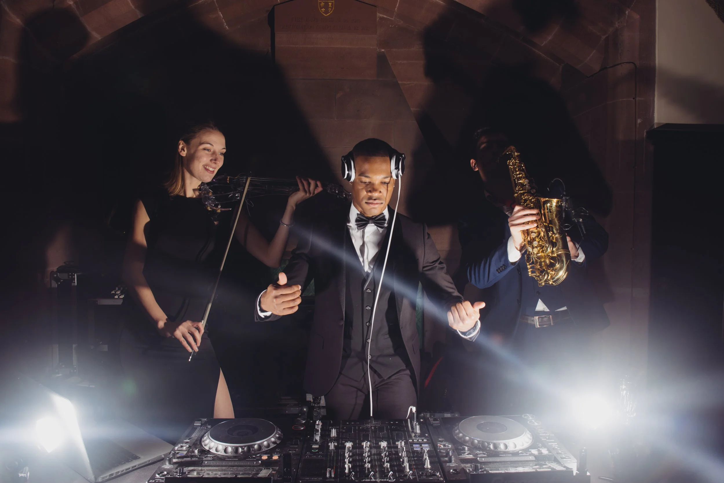 Three people, including a DJ wearing headphones, dressed in formal attire with tuxedos, are performing at a party or event. One woman is playing the violin, and a man is playing the saxophone. They are standing behind DJ equipment on a stage with wooden wall paneling in the background. Bright light shines from below, creating a dramatic atmosphere.