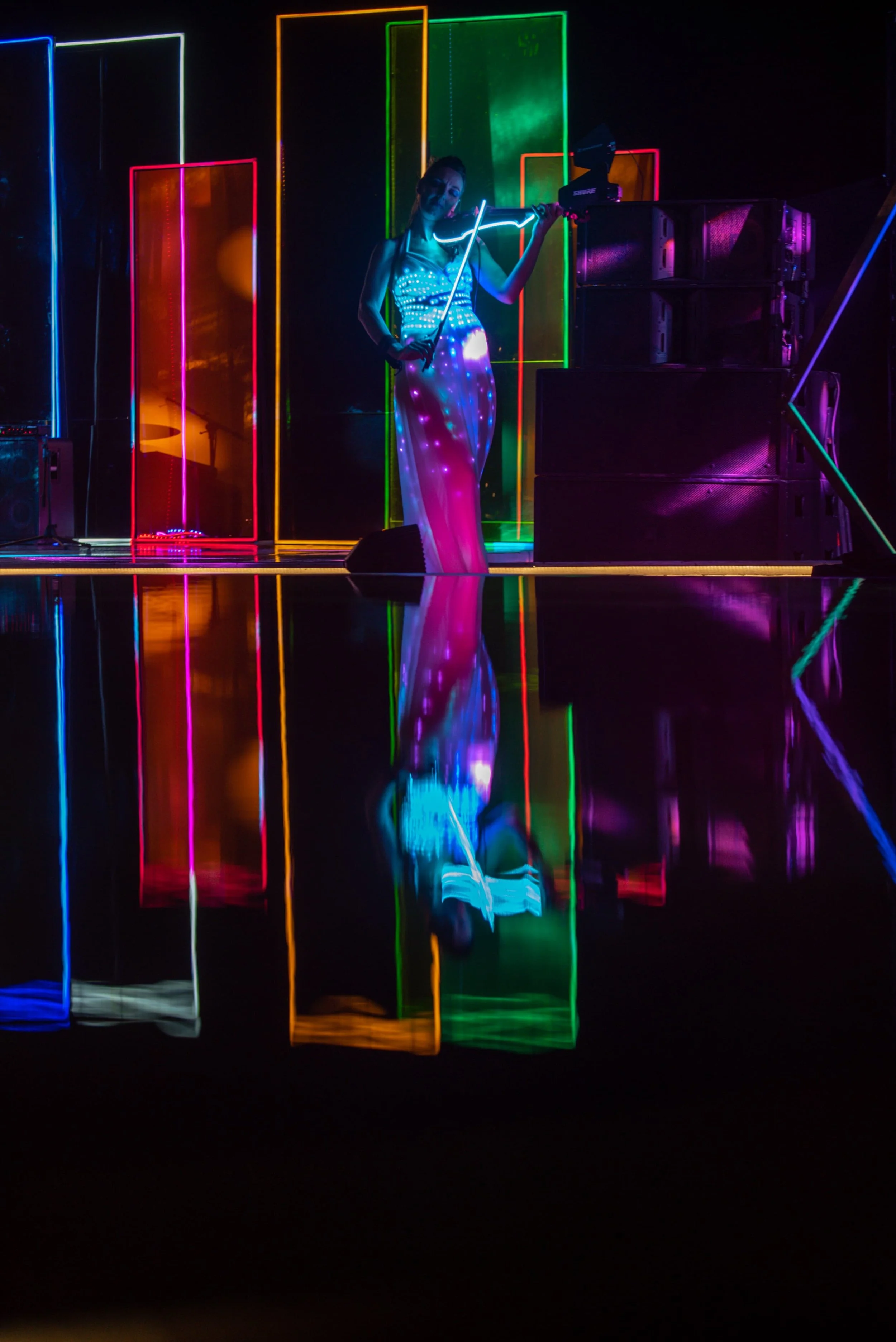 A woman dressed in a long, flowing dress made of LED lights holding and playing a violin in front of a colorful neon-lit stage with vertical light panels.