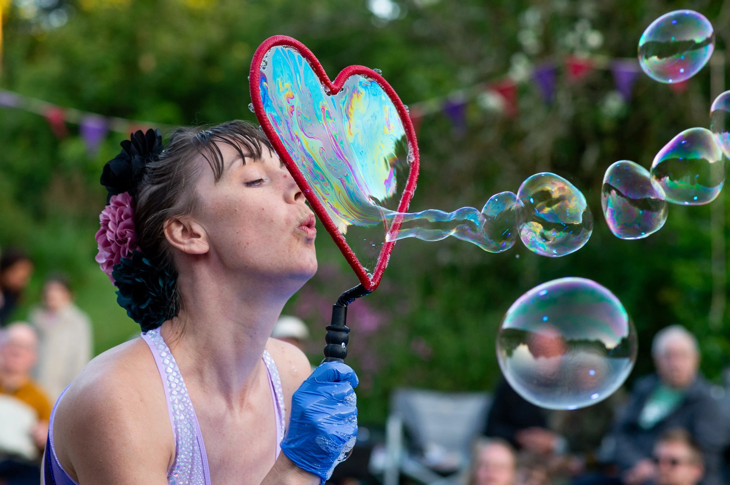 A young woman wearing a purple dress and gloves blowing soap bubbles through a heart-shaped wand at an outdoor event, with people and colorful decorations in the background.
