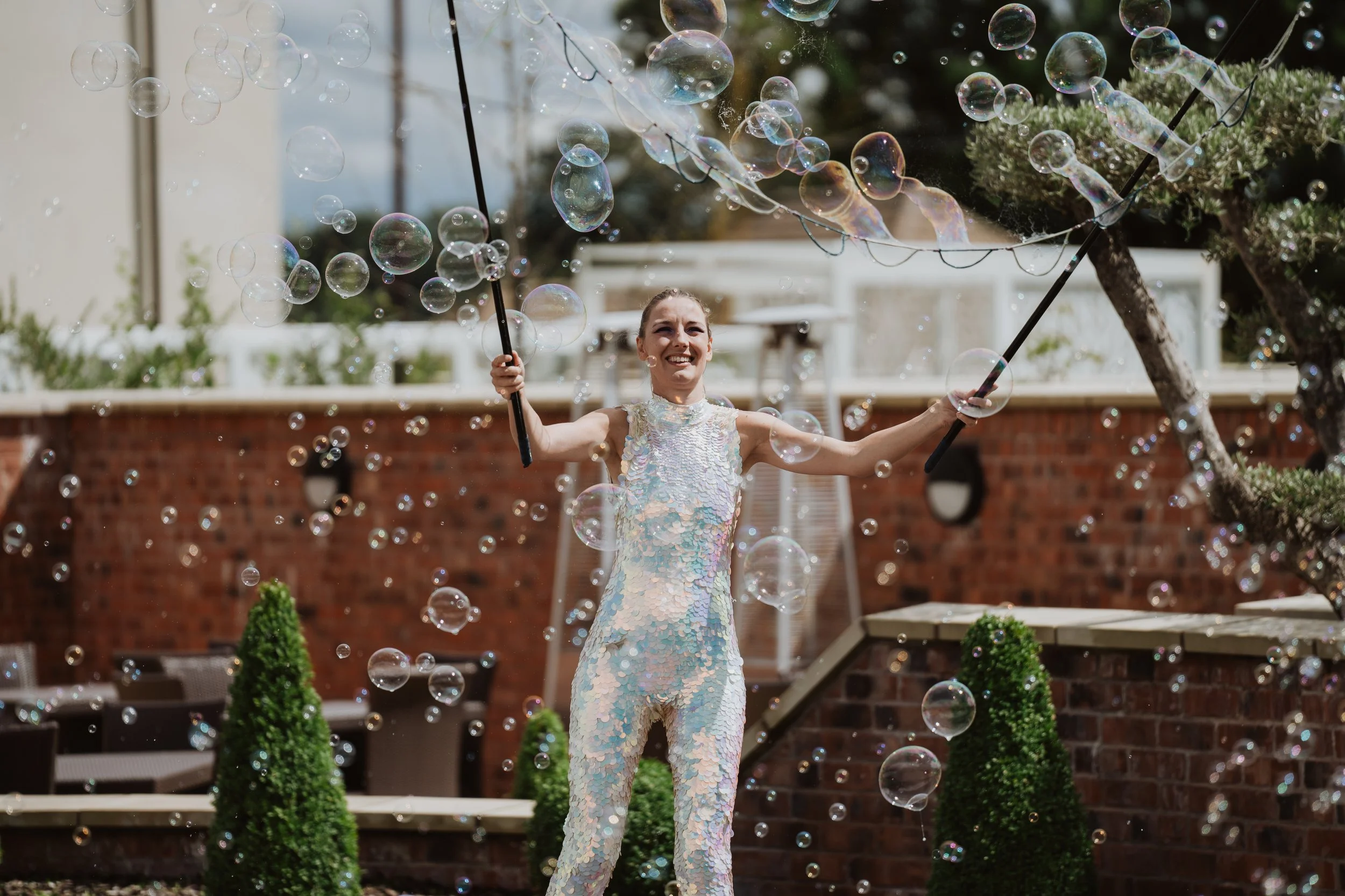 A young woman with light hair in an iridescent dress is holding two bubble wands and surrounded by floating soap bubbles in an outdoor space with brick wall and greenery.