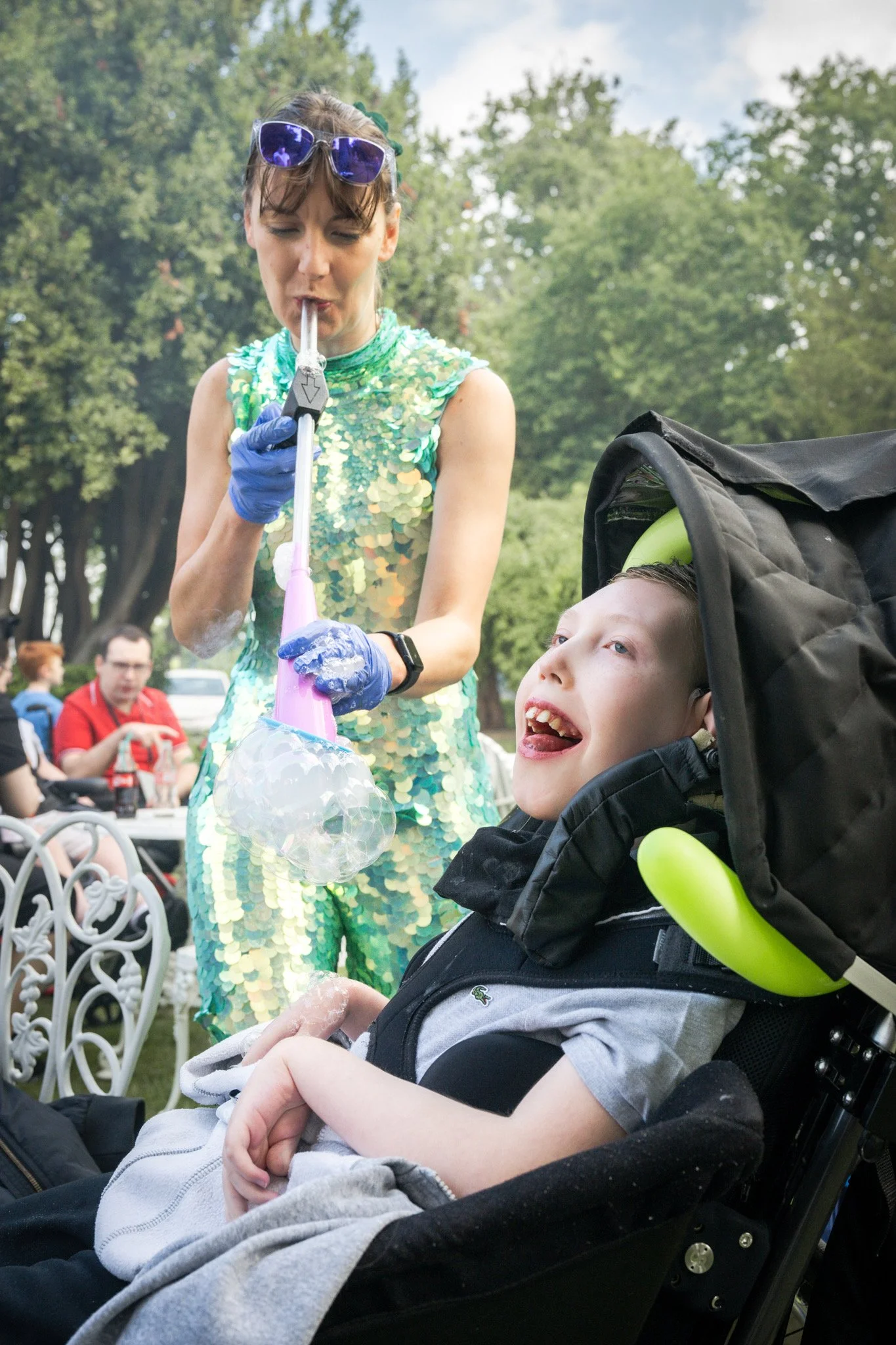 A woman with sunglasses on her head and wearing a green sequin dress blowing bubbles while a young boy in a stroller with gray shirt and gray hoodie interacts with her, outdoors in a park with trees and people in the background.