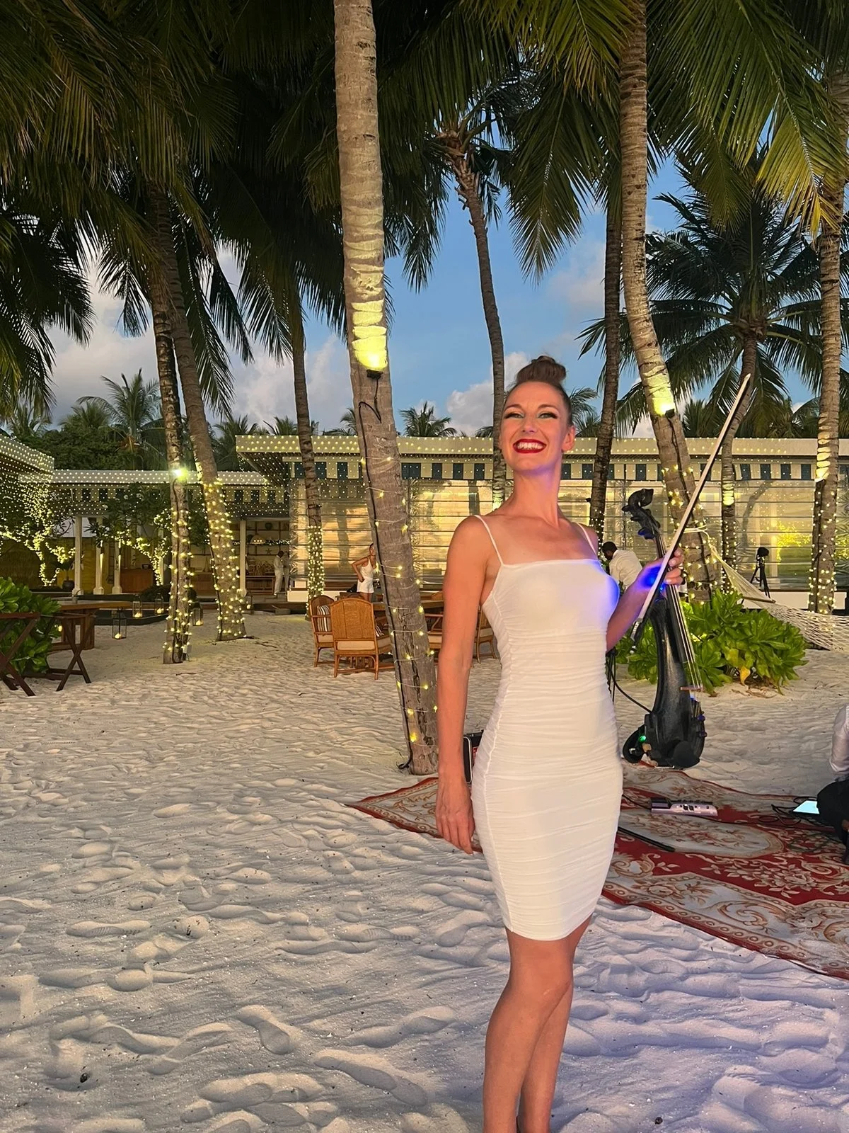 A woman in a white dress holding a violin and bow, smiling on a sandy beach with palm trees and decorated with string lights at dusk.