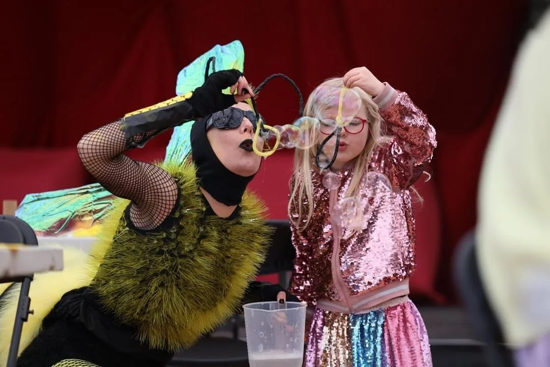 A woman dressed as a bee blowing bubbles for a young girl dressed in a pink sequin jacket and rainbow sequin pants in front of a red curtain.