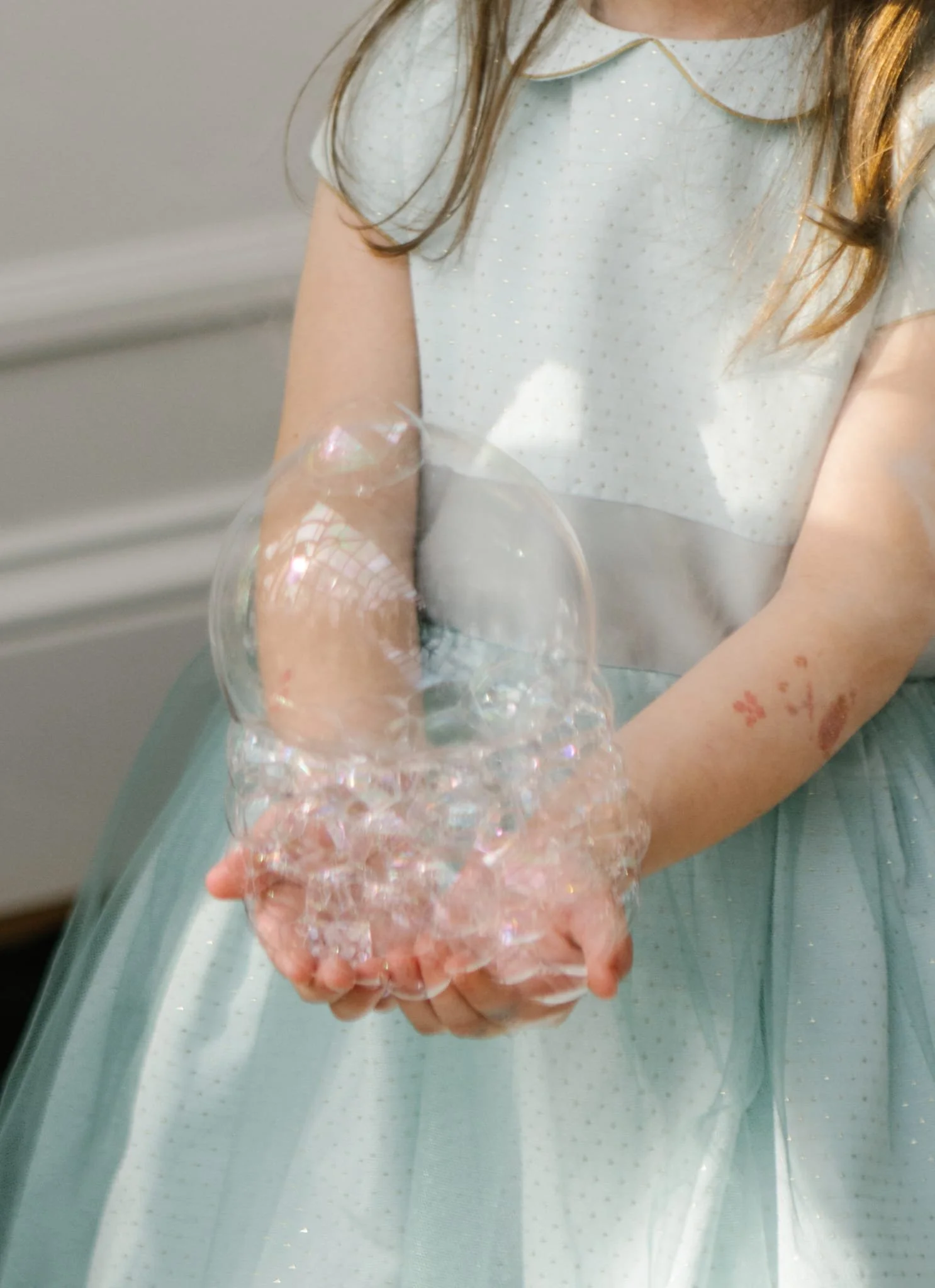 A young girl wearing a white dress with small dots and a scalloped collar is holding a large clear plastic bubble in her hands.