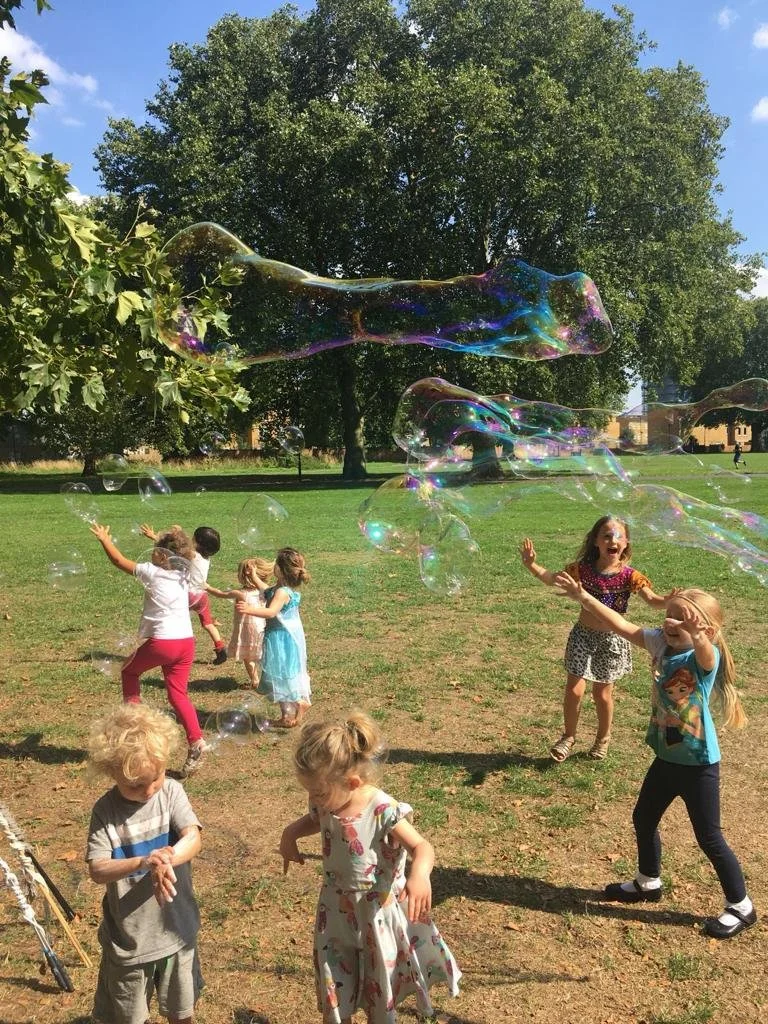 Children playing outdoors on a sunny day in a park, chasing soap bubbles near trees and a grassy field.