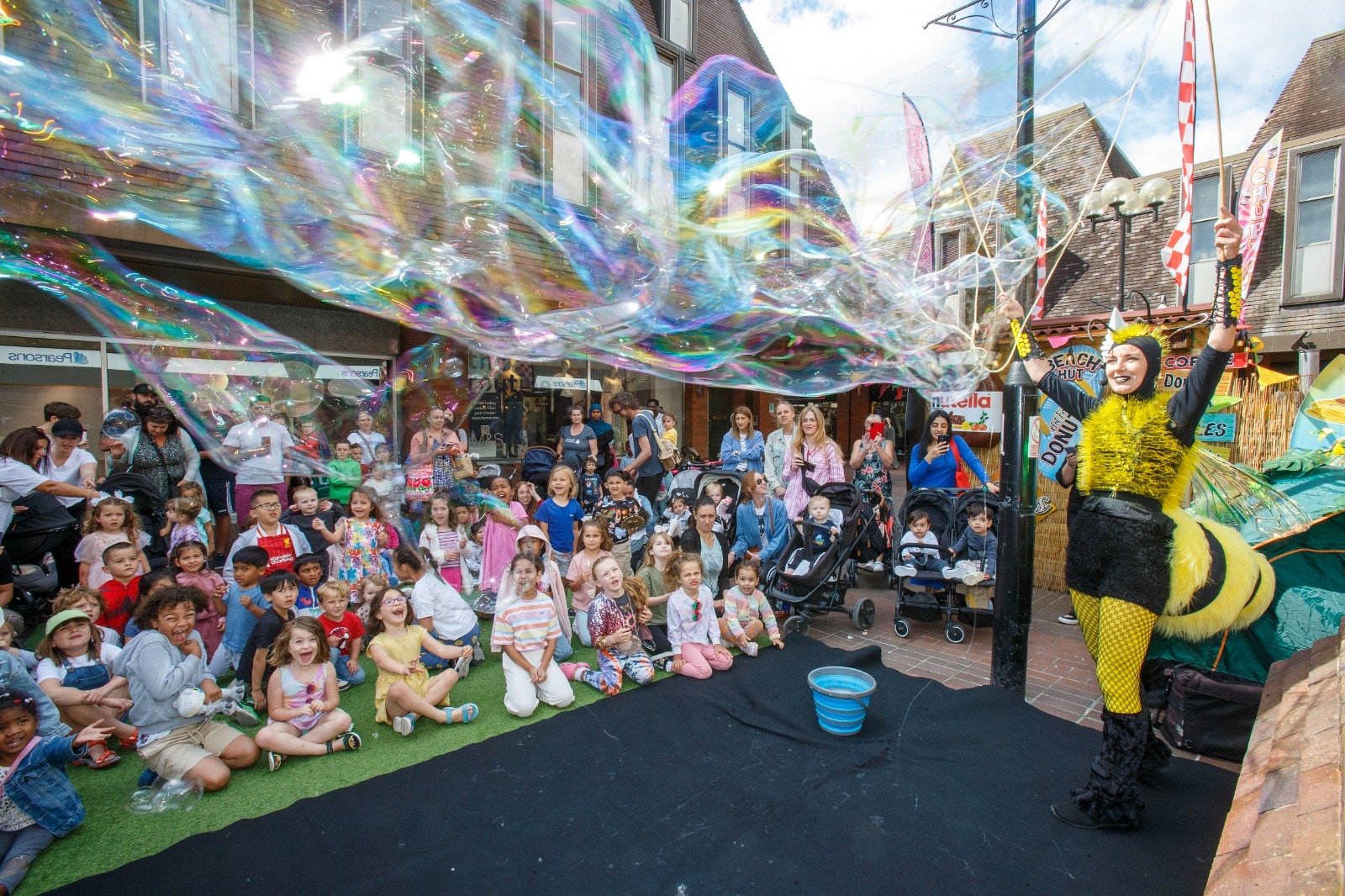 A street performer dressed as a bee entertaining a crowd with large bubbles at what appears to be a busy outdoor event or festival.
