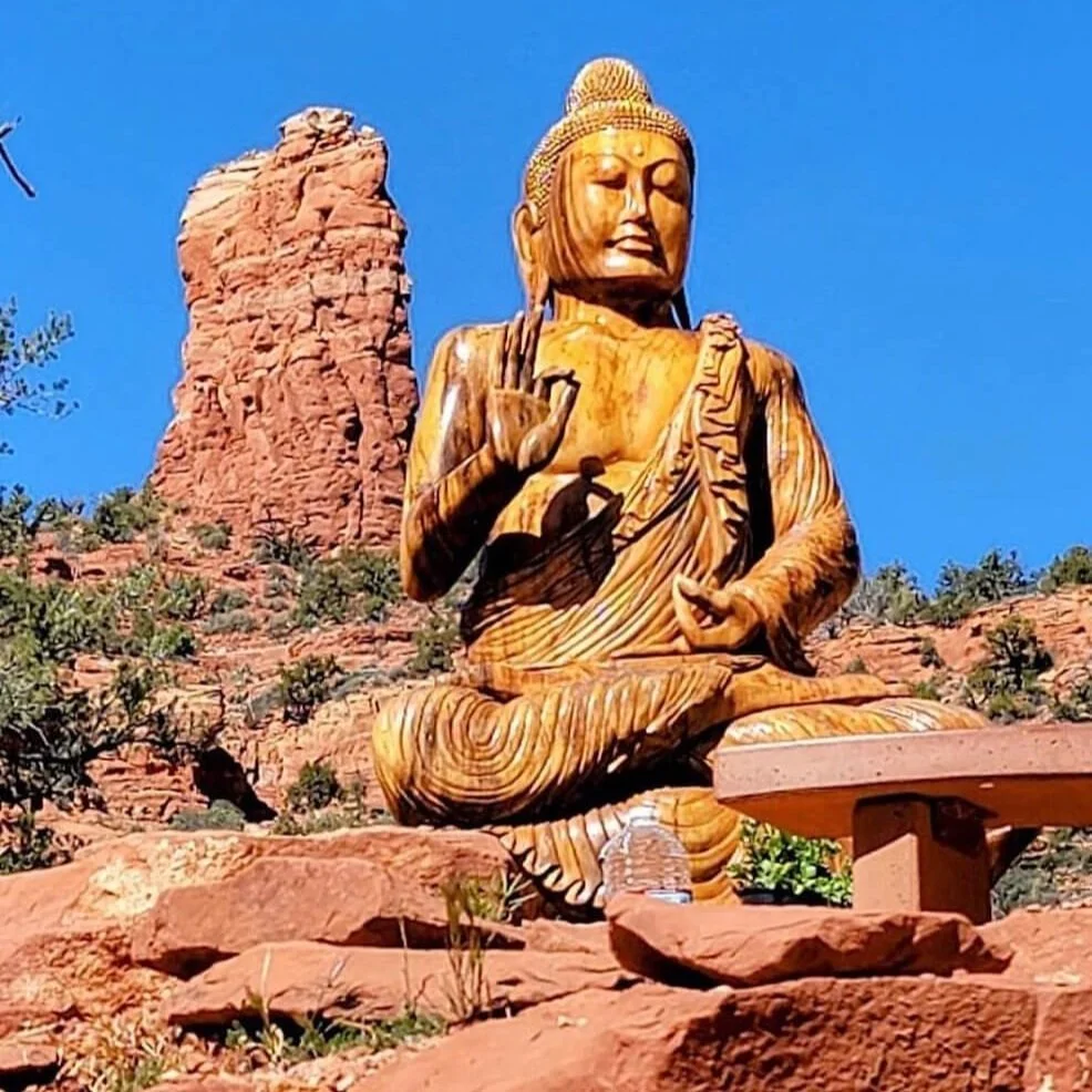 Large wooden statue of Buddha in a seated meditation pose outdoors against a red rock formation and clear blue sky.