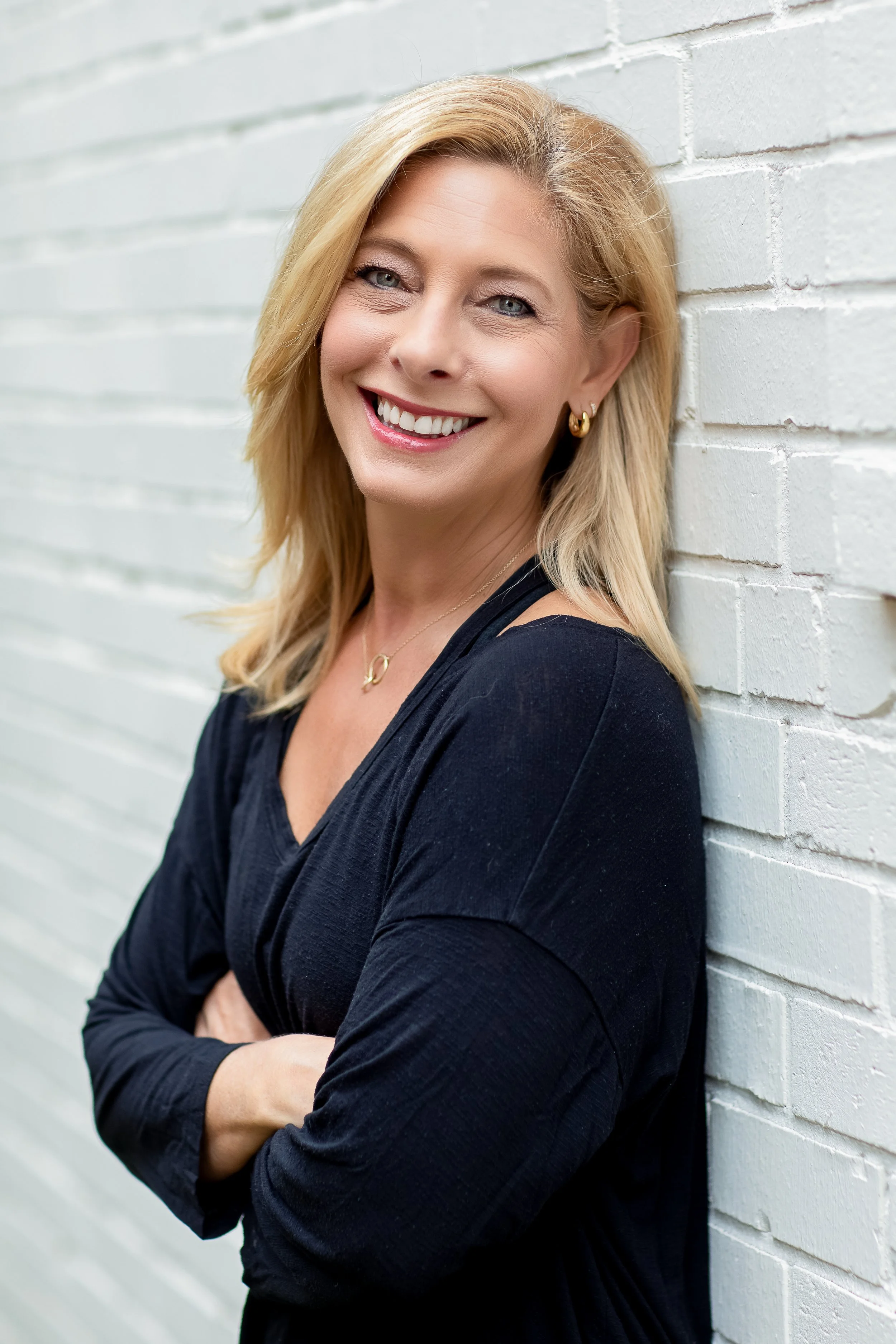 A smiling woman with blonde hair and blue eyes, wearing gold earrings and a black top, leaning against a white brick wall with her arms crossed.
