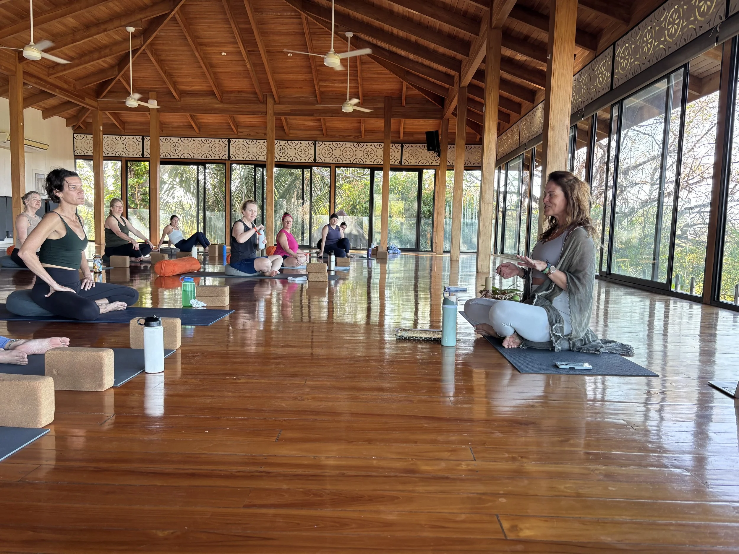A group of women participating in a yoga or meditation class in a spacious, well-lit room with large glass windows, wooden beams, and a wooden floor. The instructor is seated in front, possibly giving guidance.