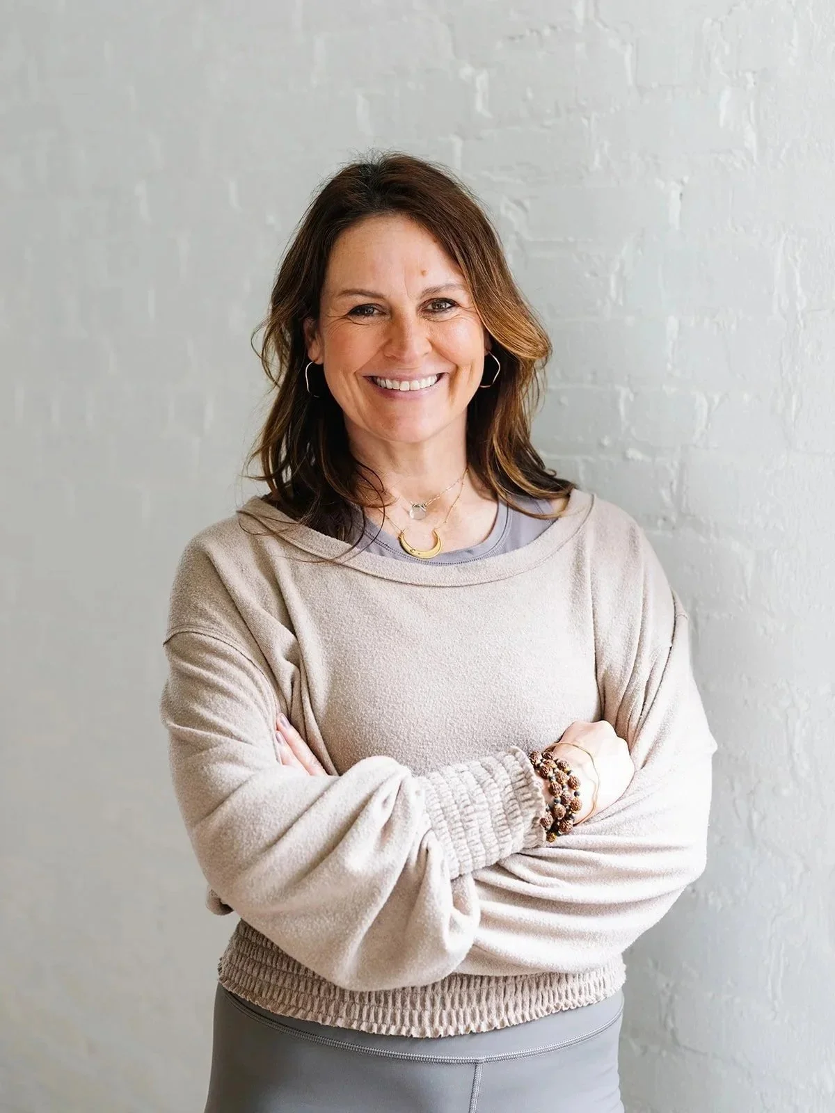 A woman with shoulder-length brown hair smiling, standing with crossed arms in front of a white brick wall, wearing a beige sweatshirt, layered necklaces, and bracelets.