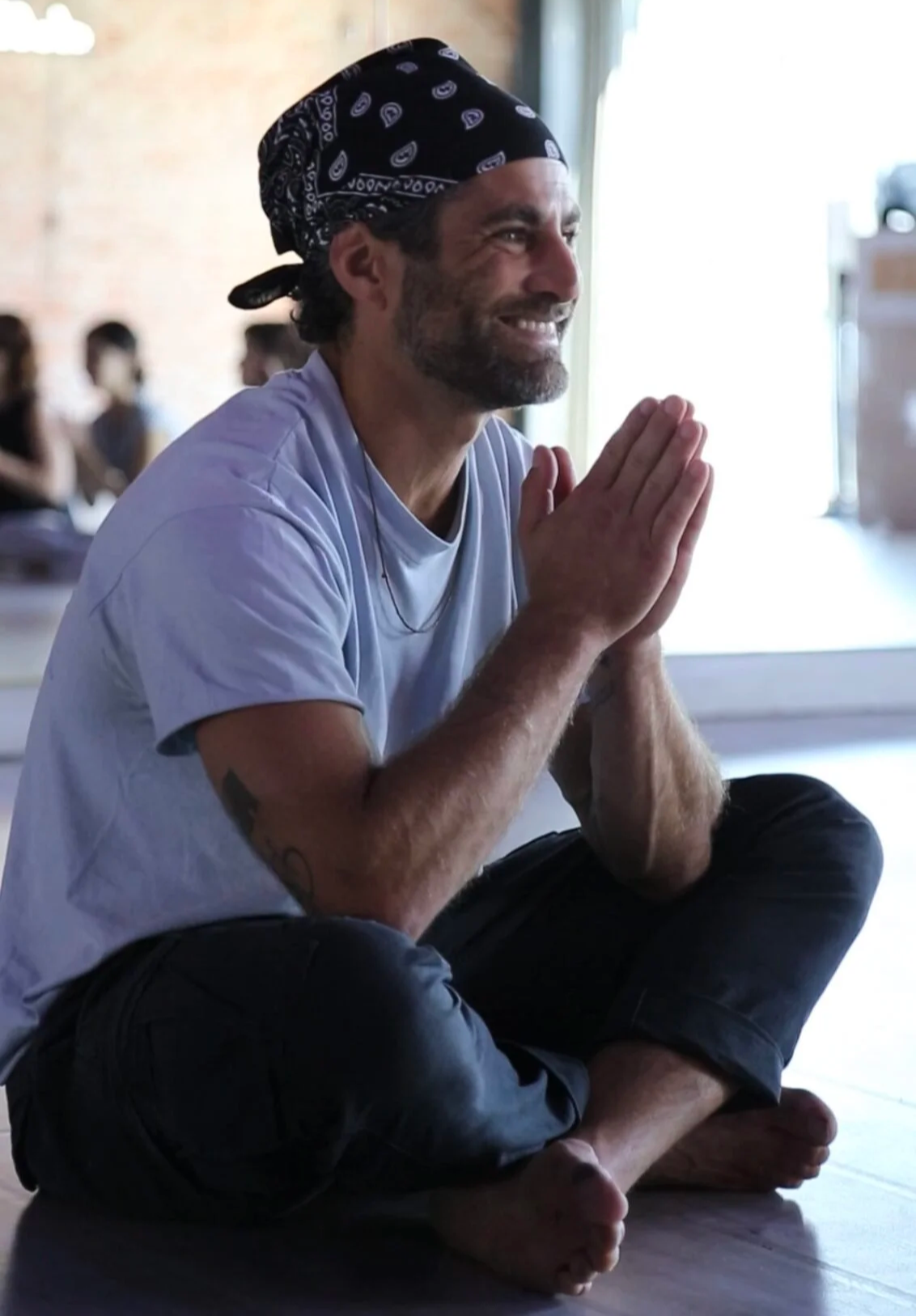 Nick Baga smiling in a bandana with hands pressed together, seated cross-legged in a dance studio