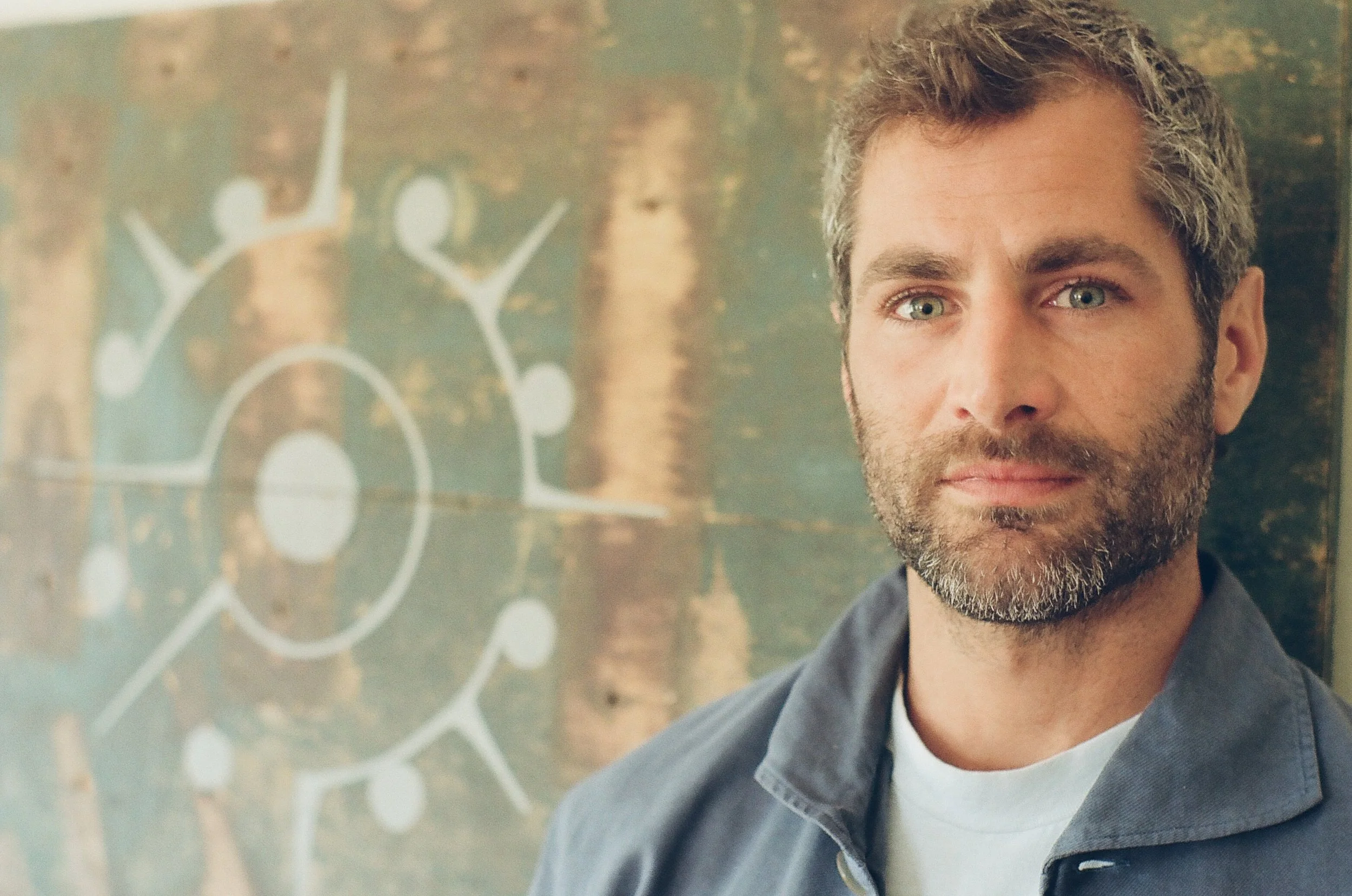 Portrait of Nick Baga in a blue work jacket in front of his own artwork