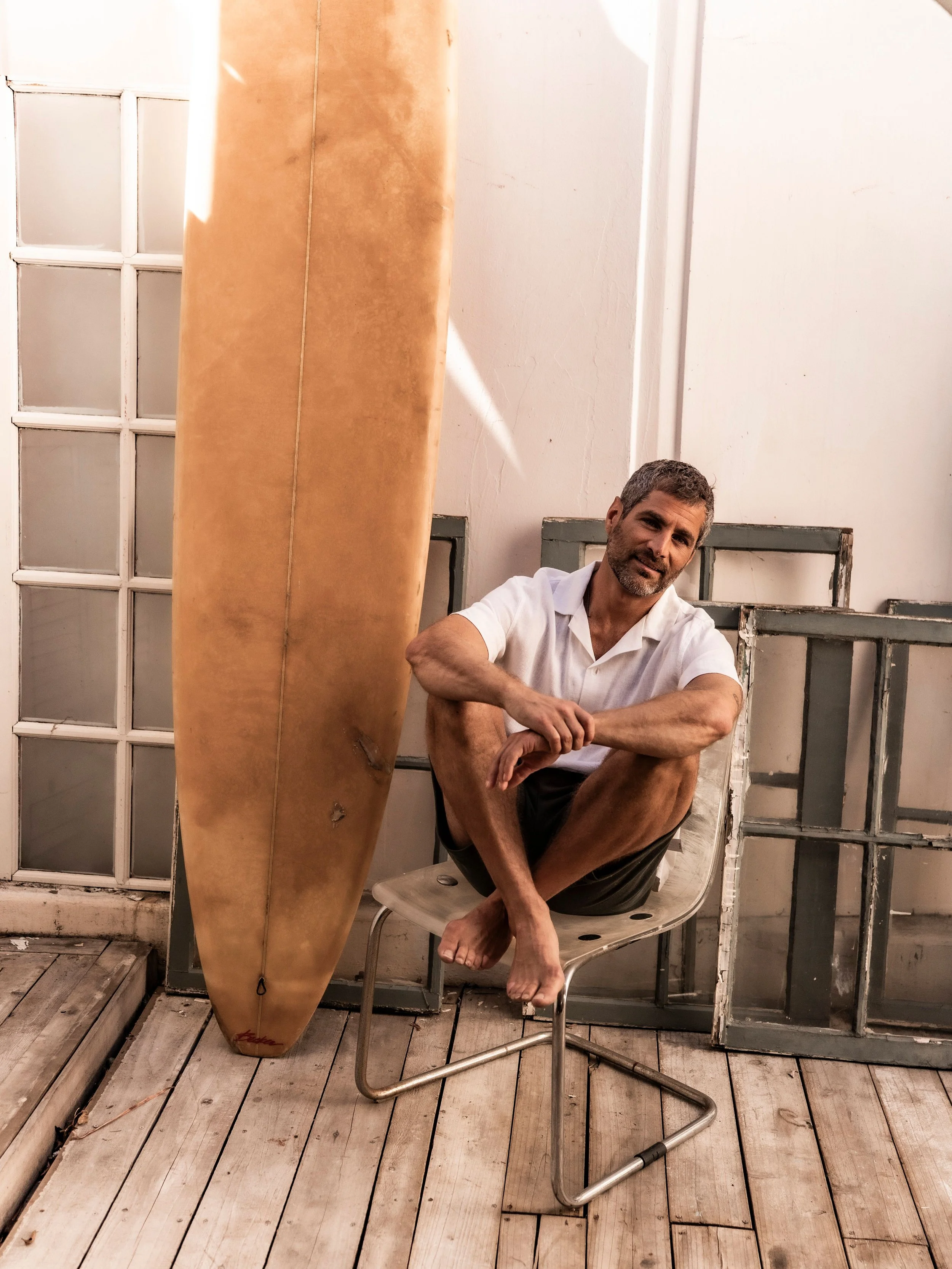 Nick Baga seated on a chair beside a vintage surfboard, arms resting on his knees