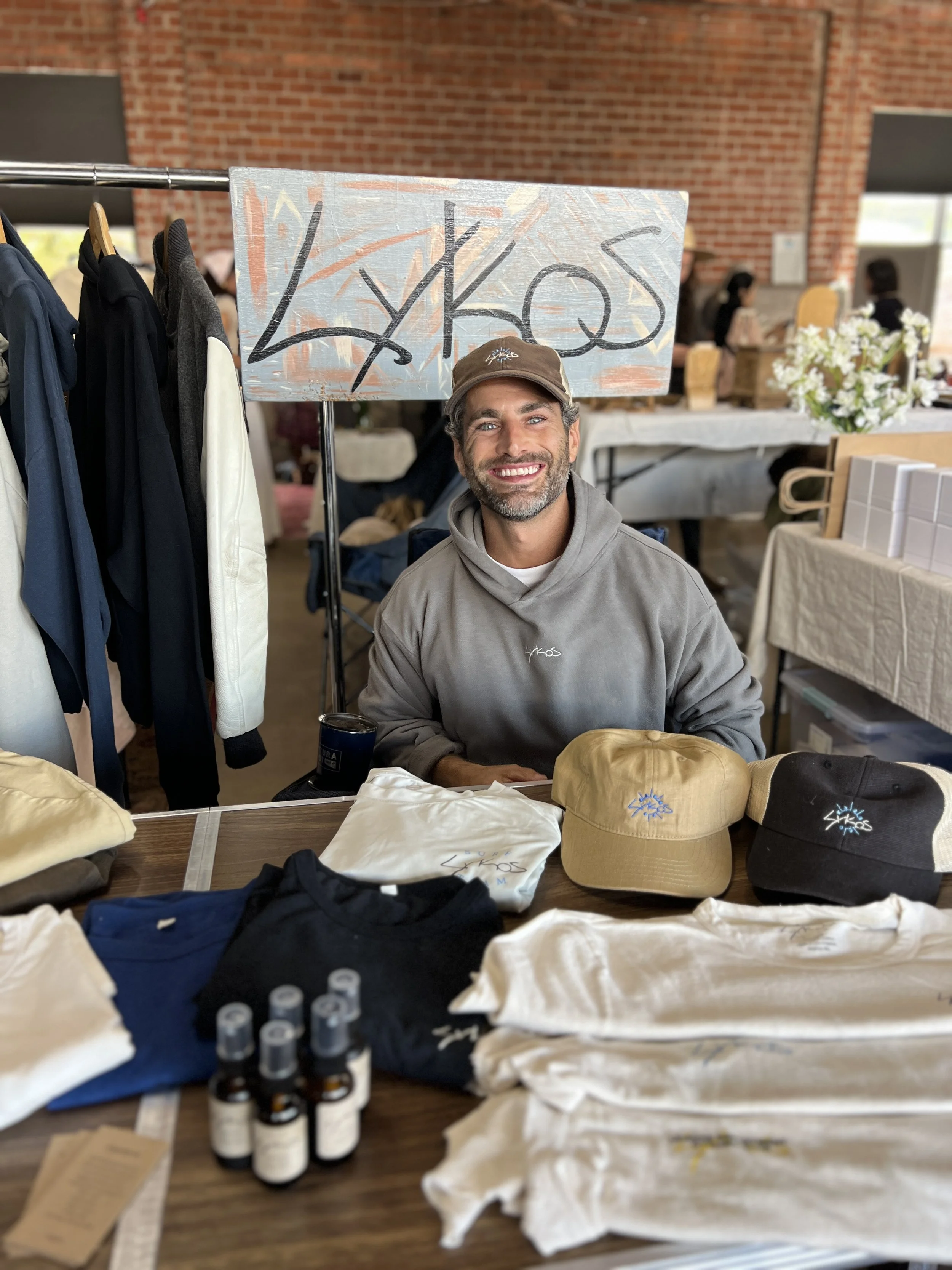 Nick Baga smiling behind a Lykos pop-up table of hemp apparel, caps and fragrance