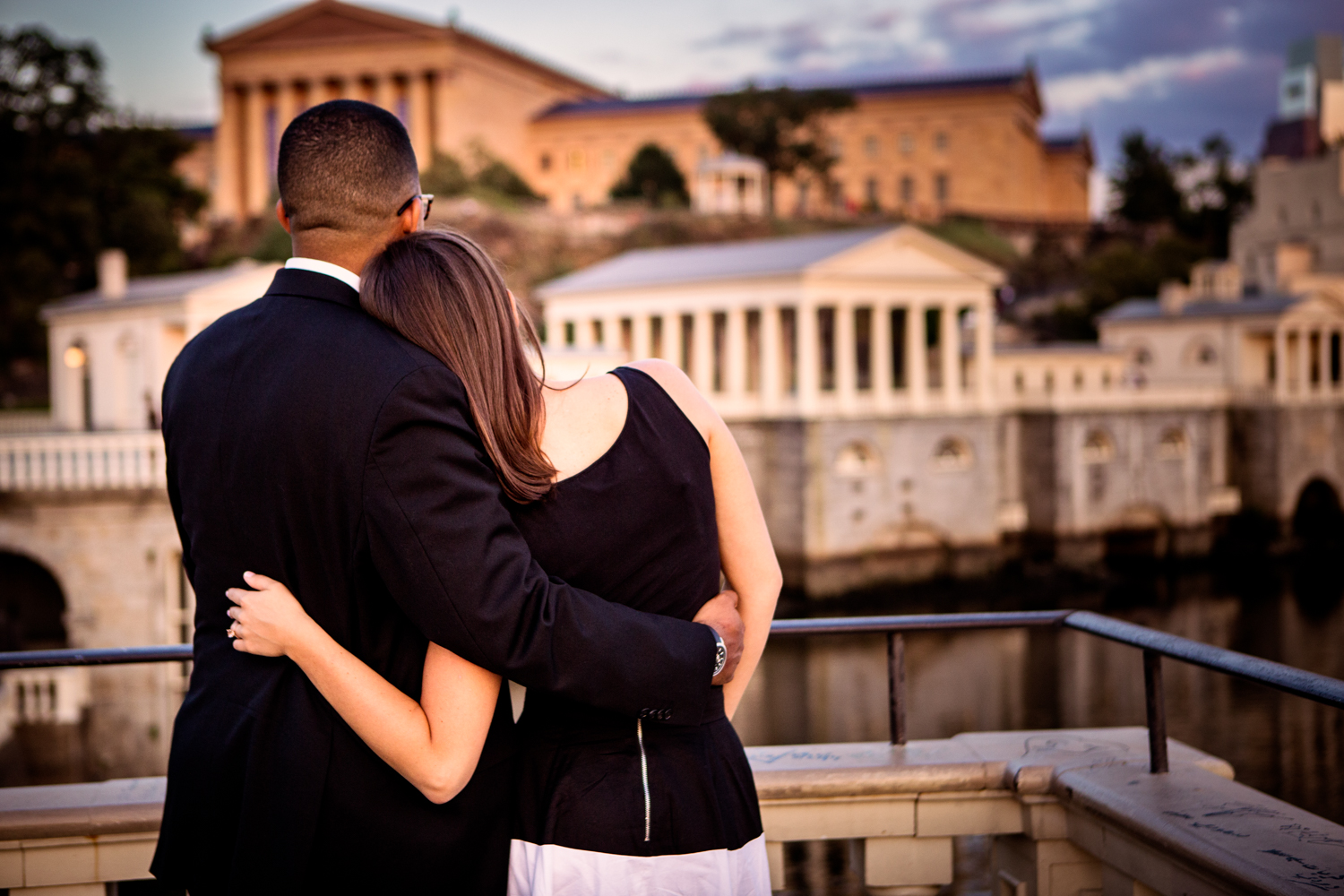 Engagement photograph of a woman leaning on her fiance's shoulder as they look at the Philadelphia Museum of Art and Waterworks