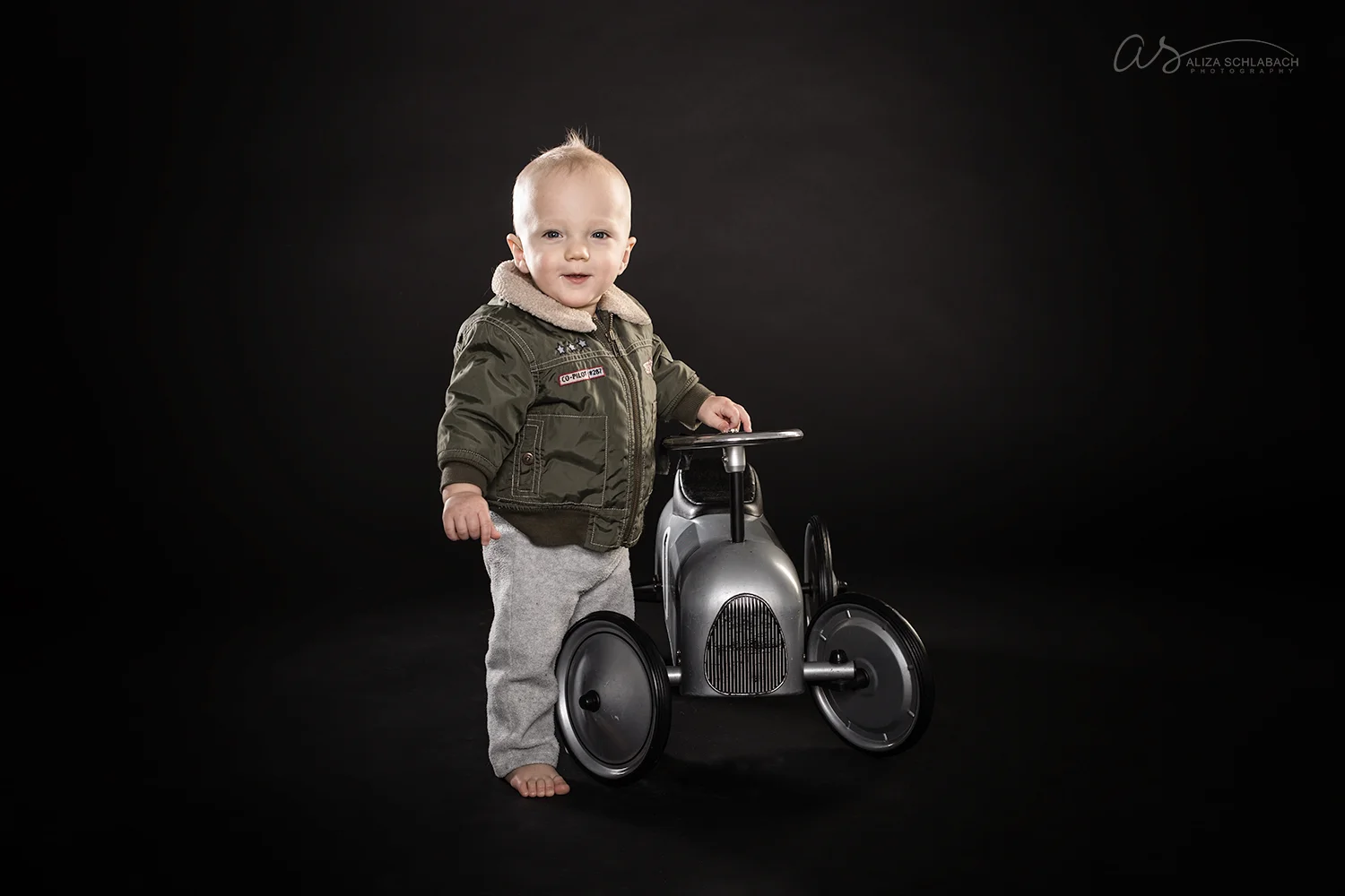 Portrait of a 1 year old baby in a bomber jacket next to a retro push car toy