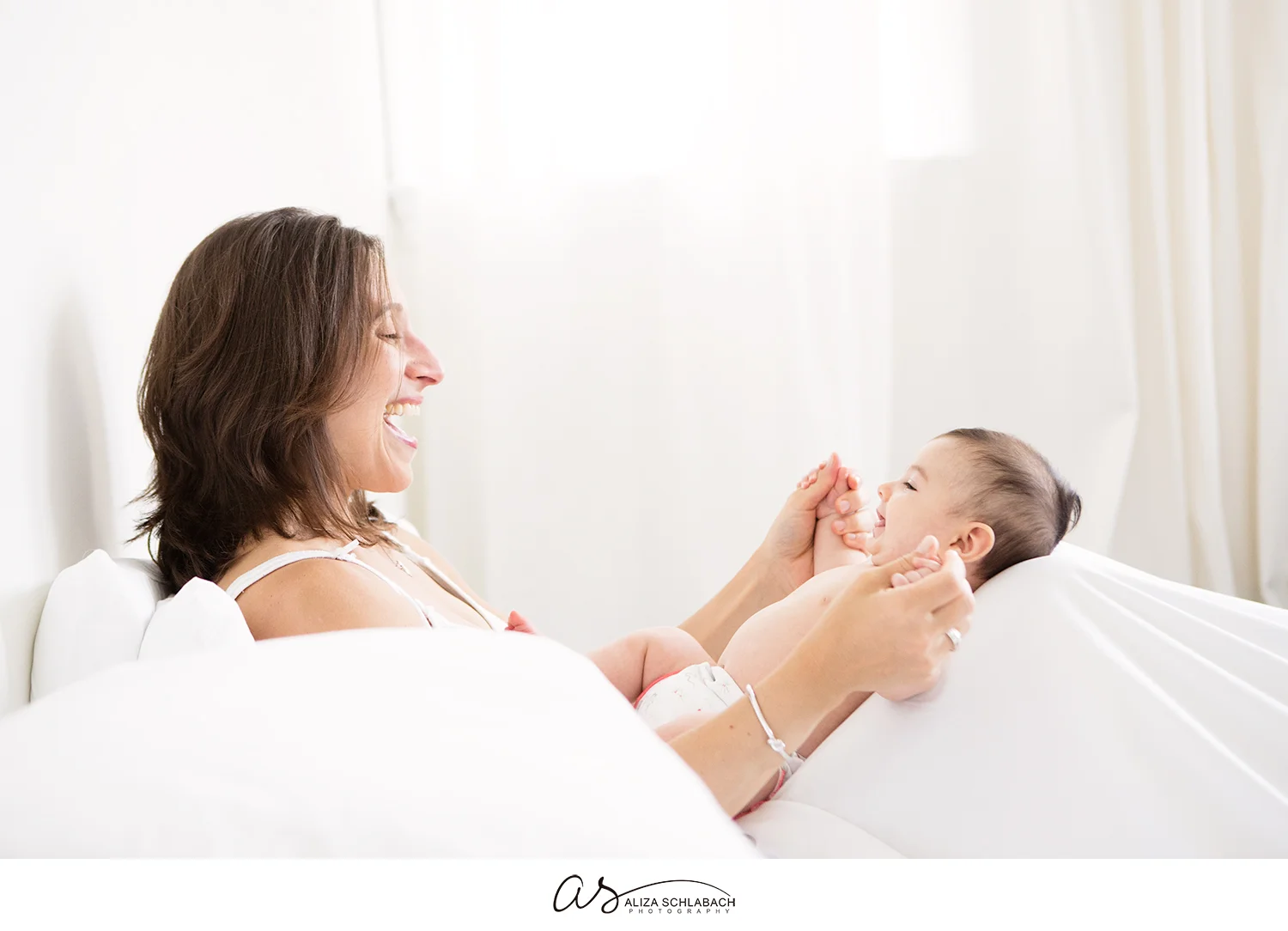 White photo of a laughing mother playing with her baby on a bed