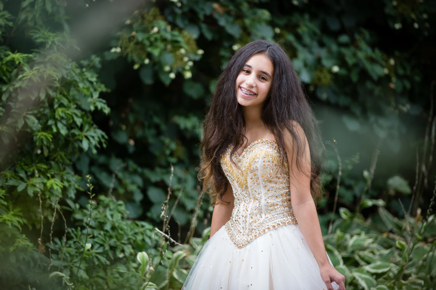 Photo of smiling bat mitzvah girl in strapless gown in front of greenery